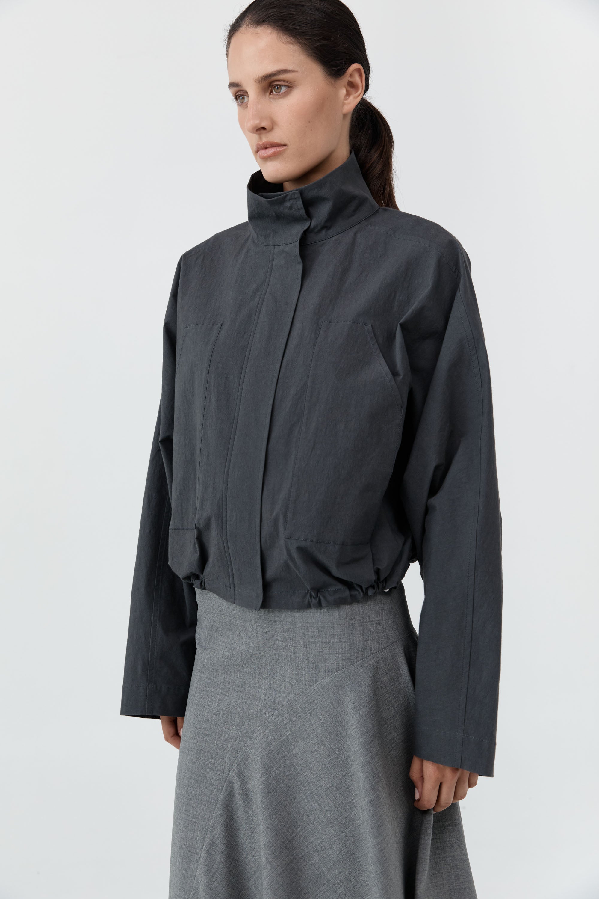 A model wearing a dark gray cropped jacket with a high mock neck and a matching gray skirt, photographed against a plain light gray studio background.