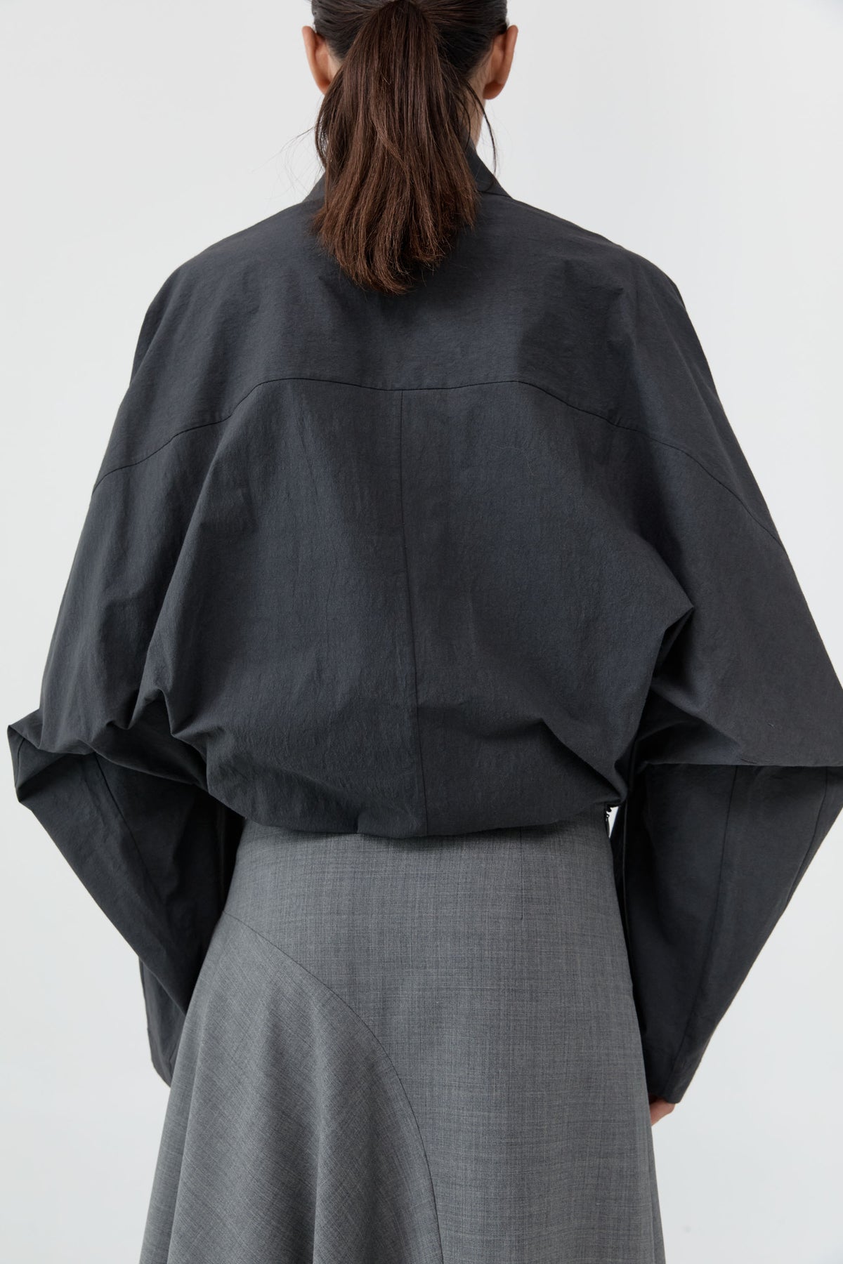 Rear view of a model wearing a dark charcoal gray oversized long-sleeve top and a textured gray skirt against a light gray studio background.
