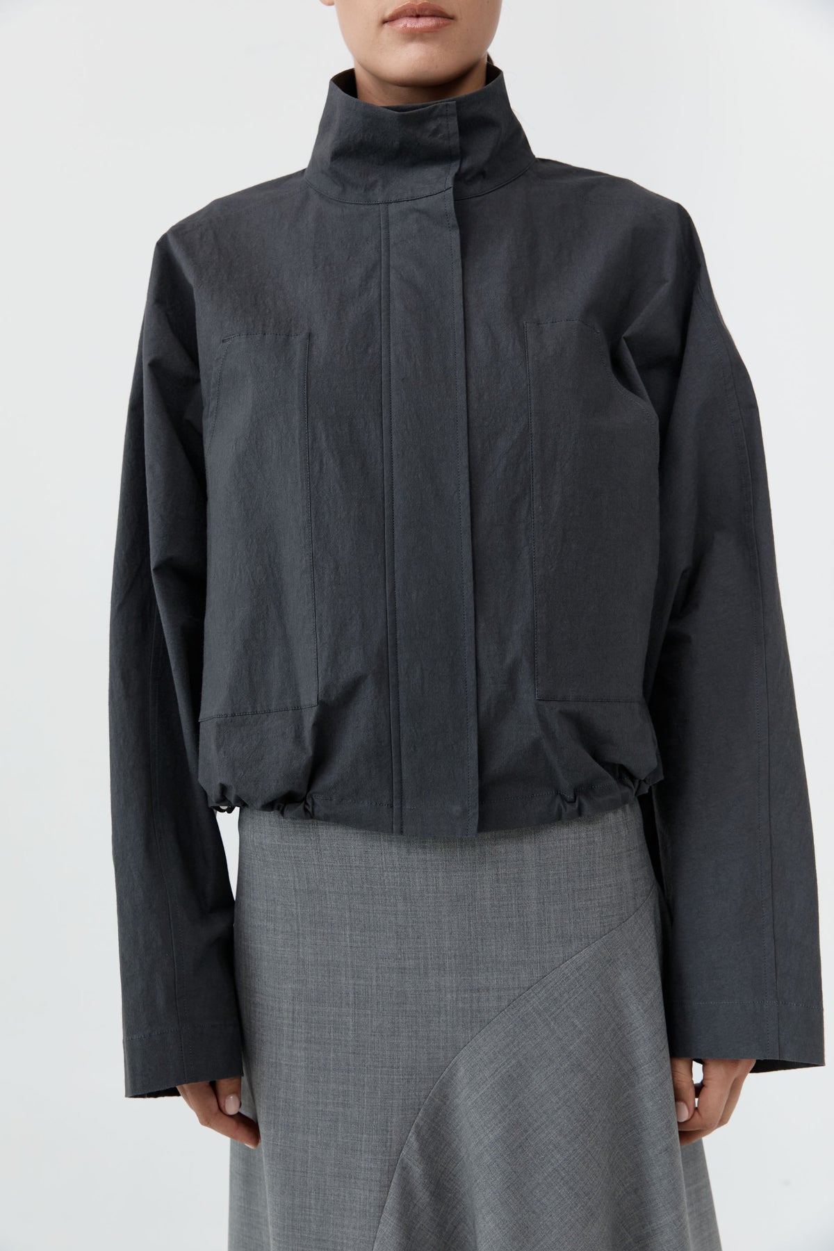 A model wearing a dark charcoal gray cropped jacket with a high mock neck collar and a gray textured skirt, shot against a plain light gray background.
