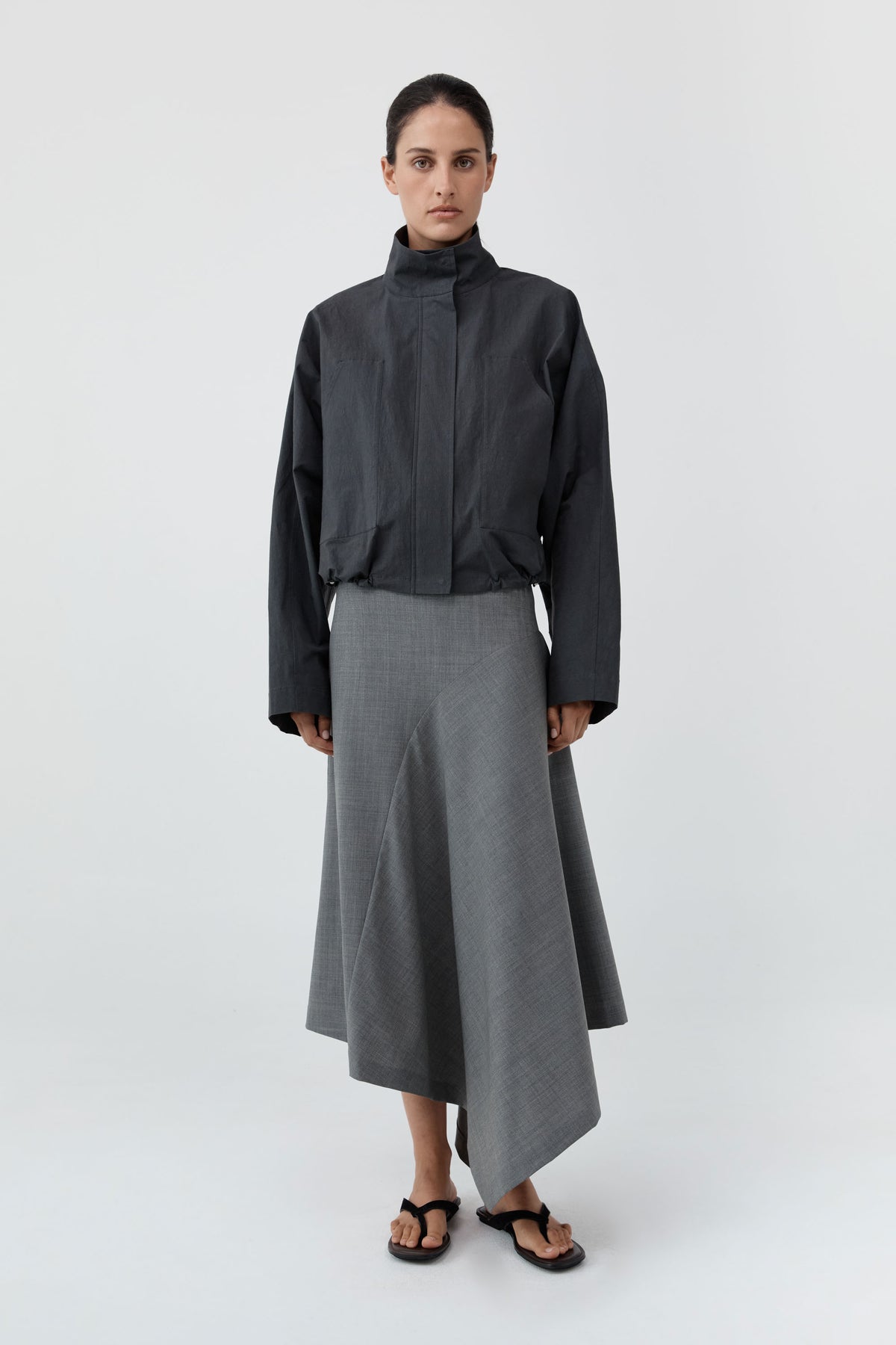 A model stands against a light gray studio background wearing a dark charcoal high-neck jacket and a gray asymmetrical midi skirt, paired with black sandals.