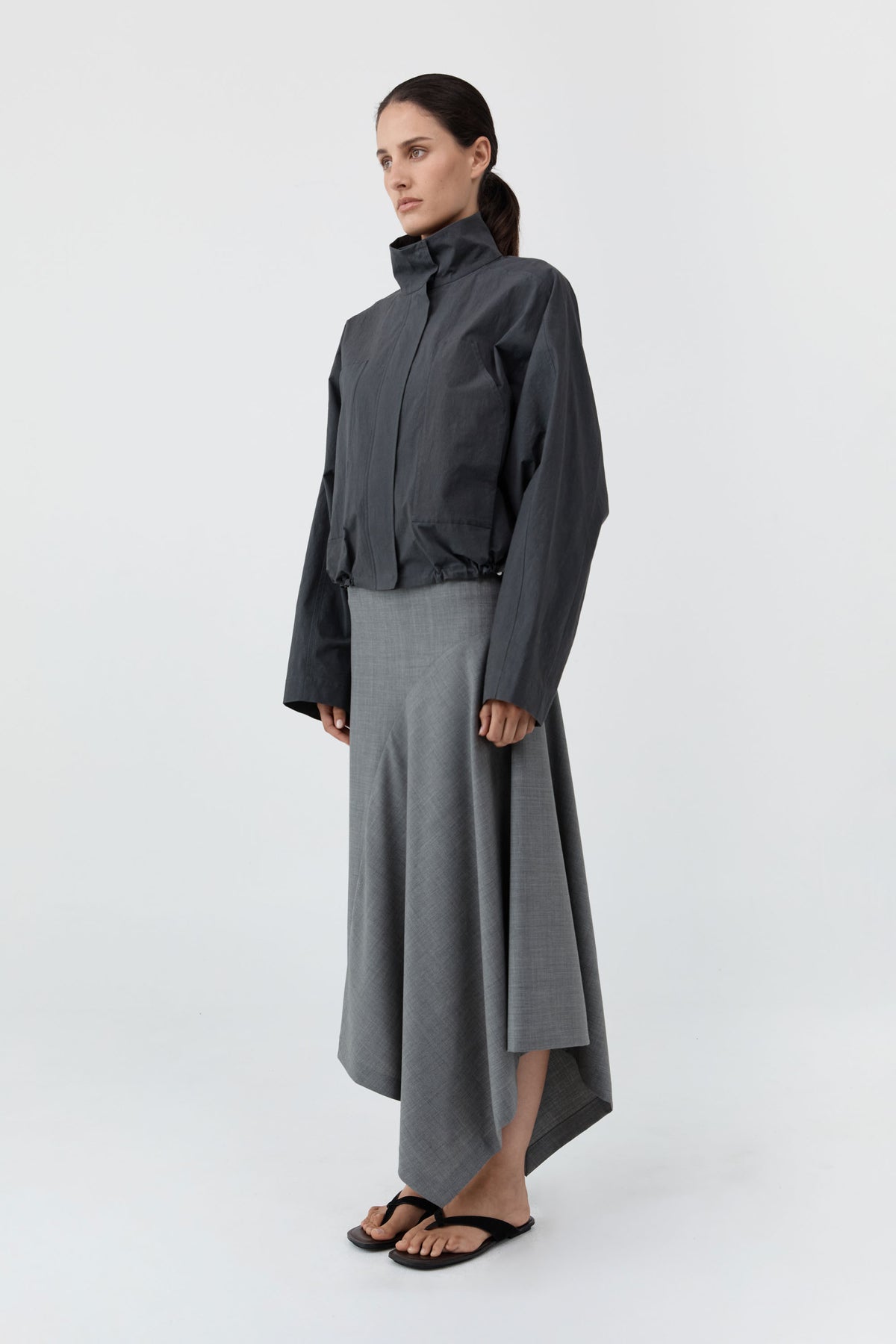 A model stands in a studio wearing a dark charcoal gray high-collar jacket over a gray high-low skirt, paired with black sandals, against a light gray background.