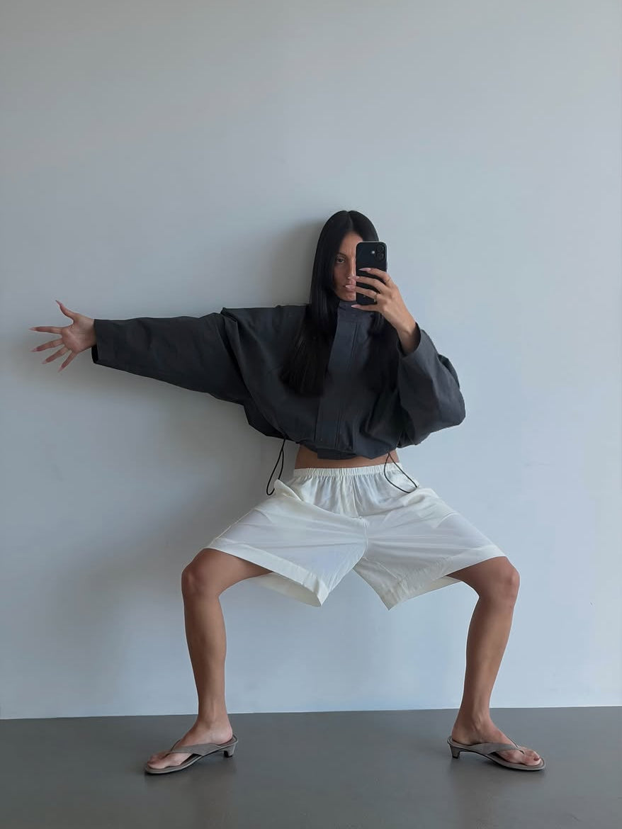 A person posing in a mirror selfie wearing a black cropped long-sleeve top, white drawstring shorts, and silver flip-flop sandals against a plain light gray wall.