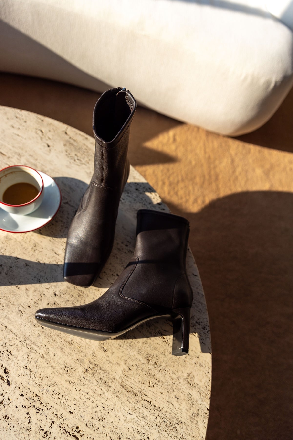 A Emery Torres Boot in bistre stretch leather, one upright and one on its side on a stone table beside an espresso cup