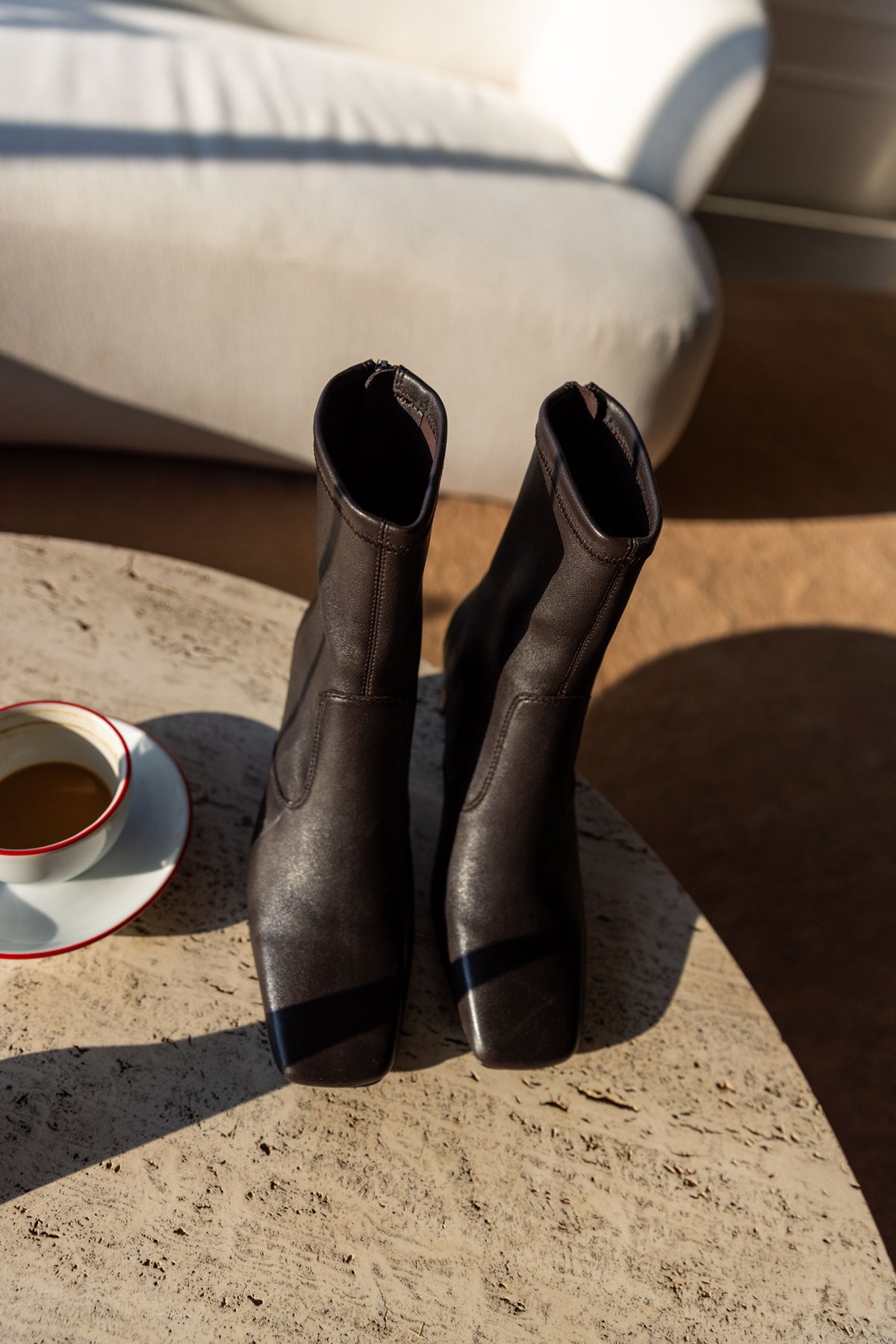 A Emery Torres Boot in bistre stretch leather, the pair standing on a sunlit stone table beside an espresso cup
