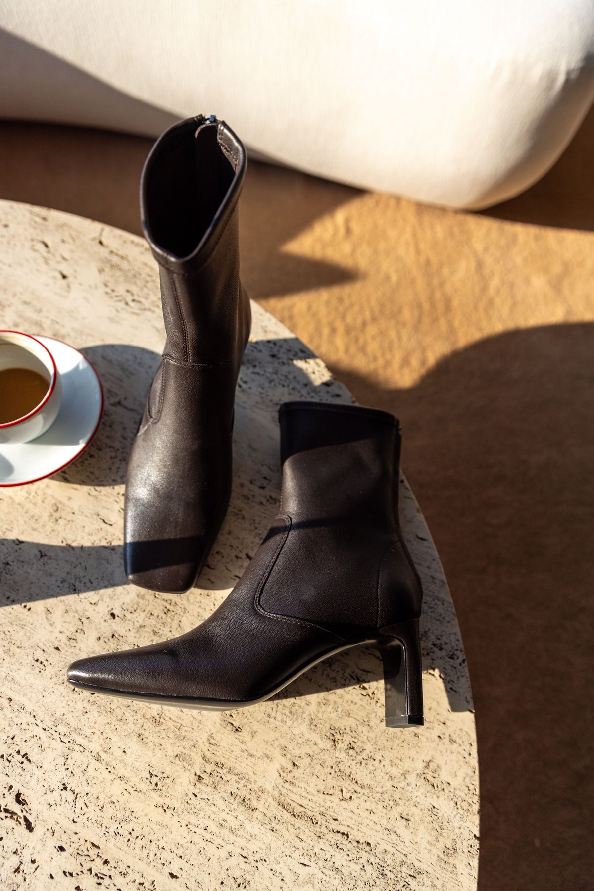 A Emery Torres Boot in bistre stretch leather, sunlit still life on stone with one boot laid on its side