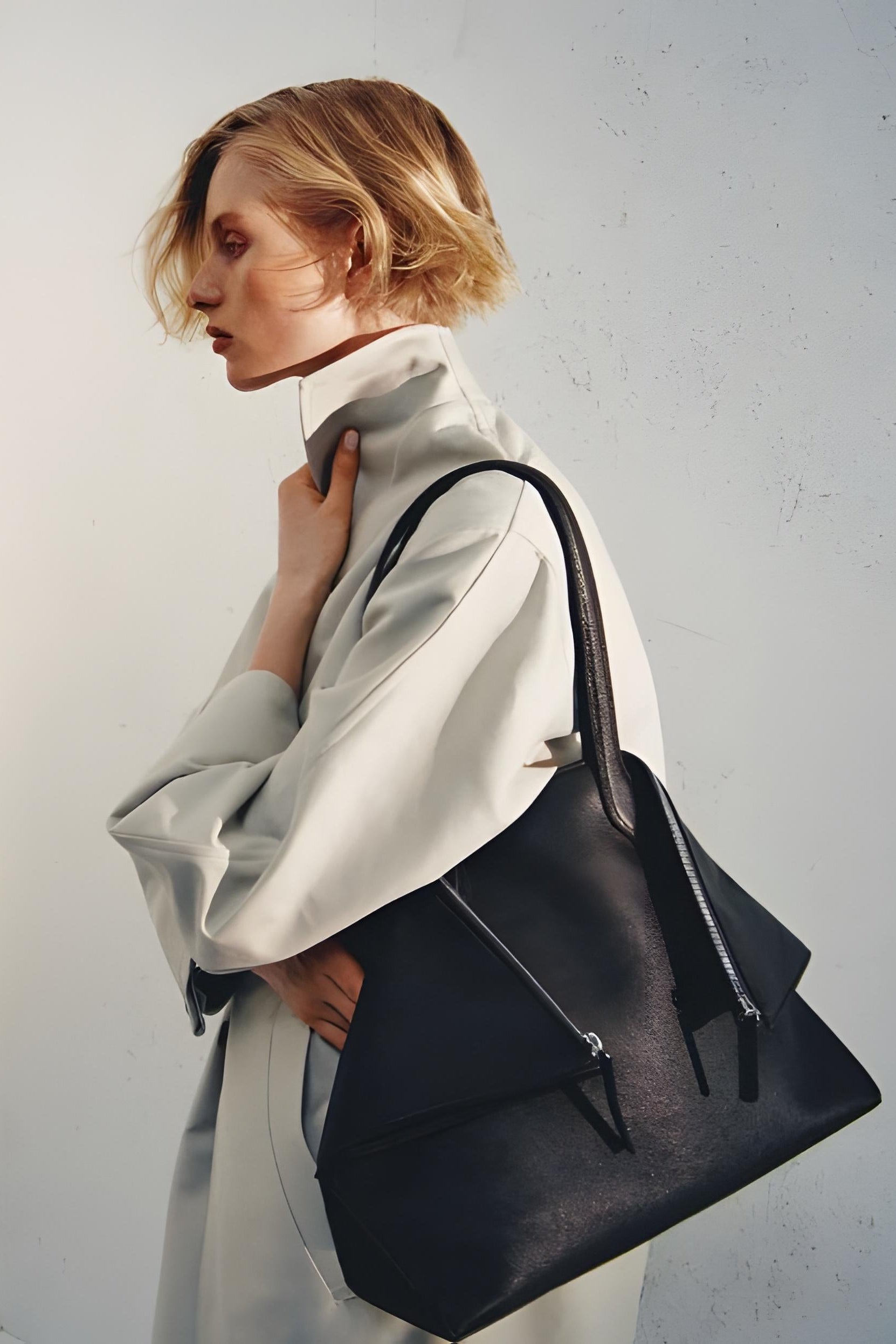 A model in a light beige trench coat carrying the Aesther Ekme Fold Bag, set against a white wall.