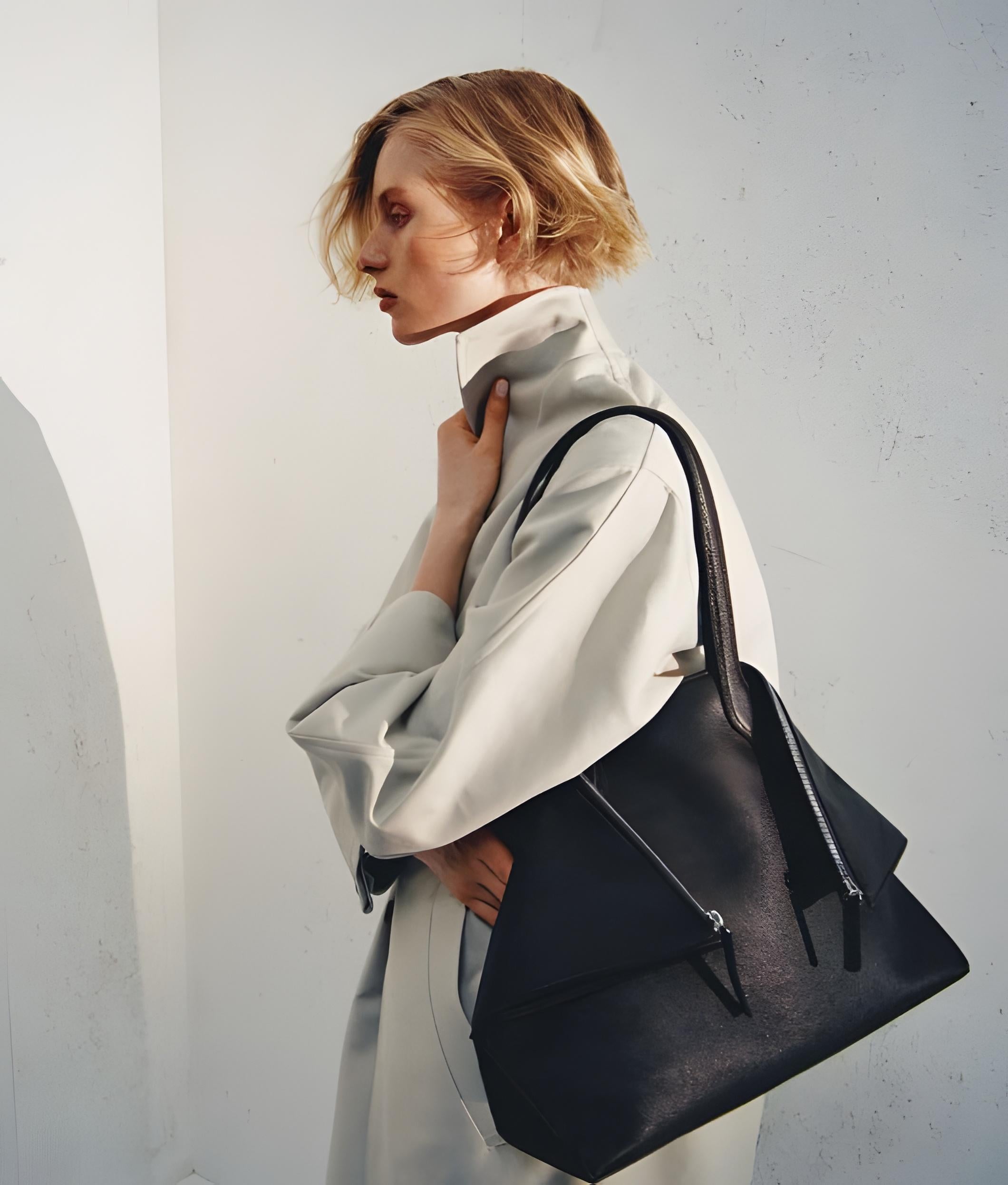 A model in a light beige trench coat carries a large black leather shoulder bag with a structured shape and double zipper details, set against a textured light gray wall.