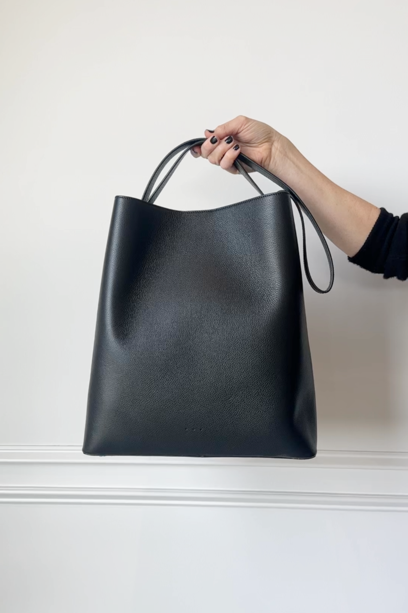 Black leather tote bag held by a hand against a white background