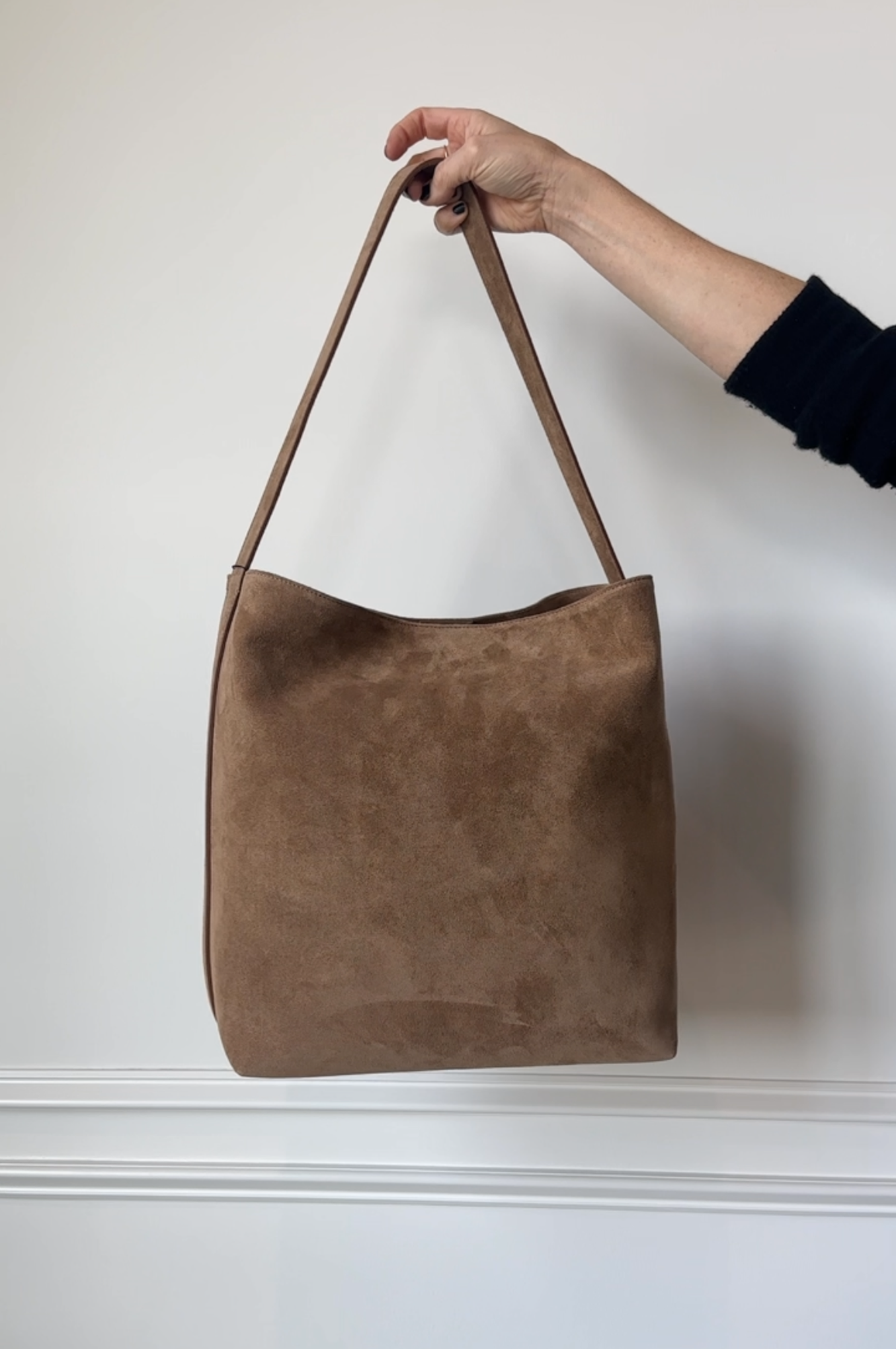 Brown suede handbag held by a hand against a white wall