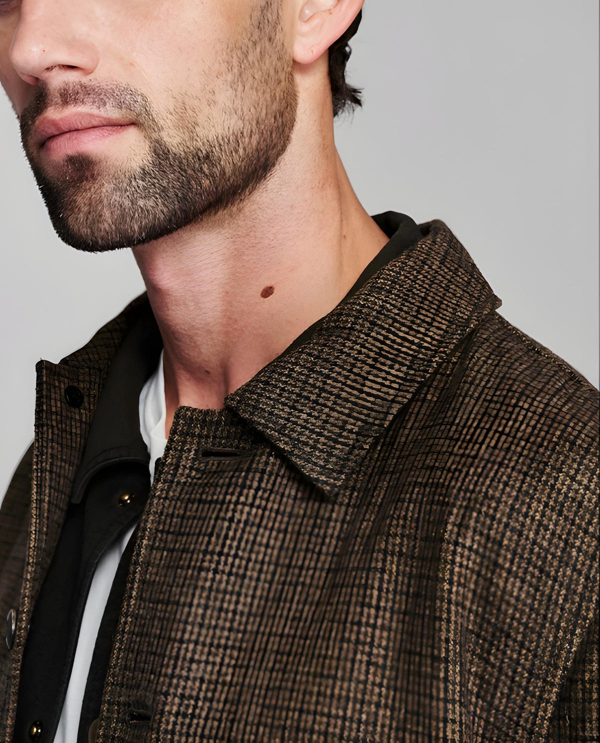 Close-up of a man wearing a brown and black micro-check patterned overshirt over a black shirt and white undershirt, against a plain gray background.