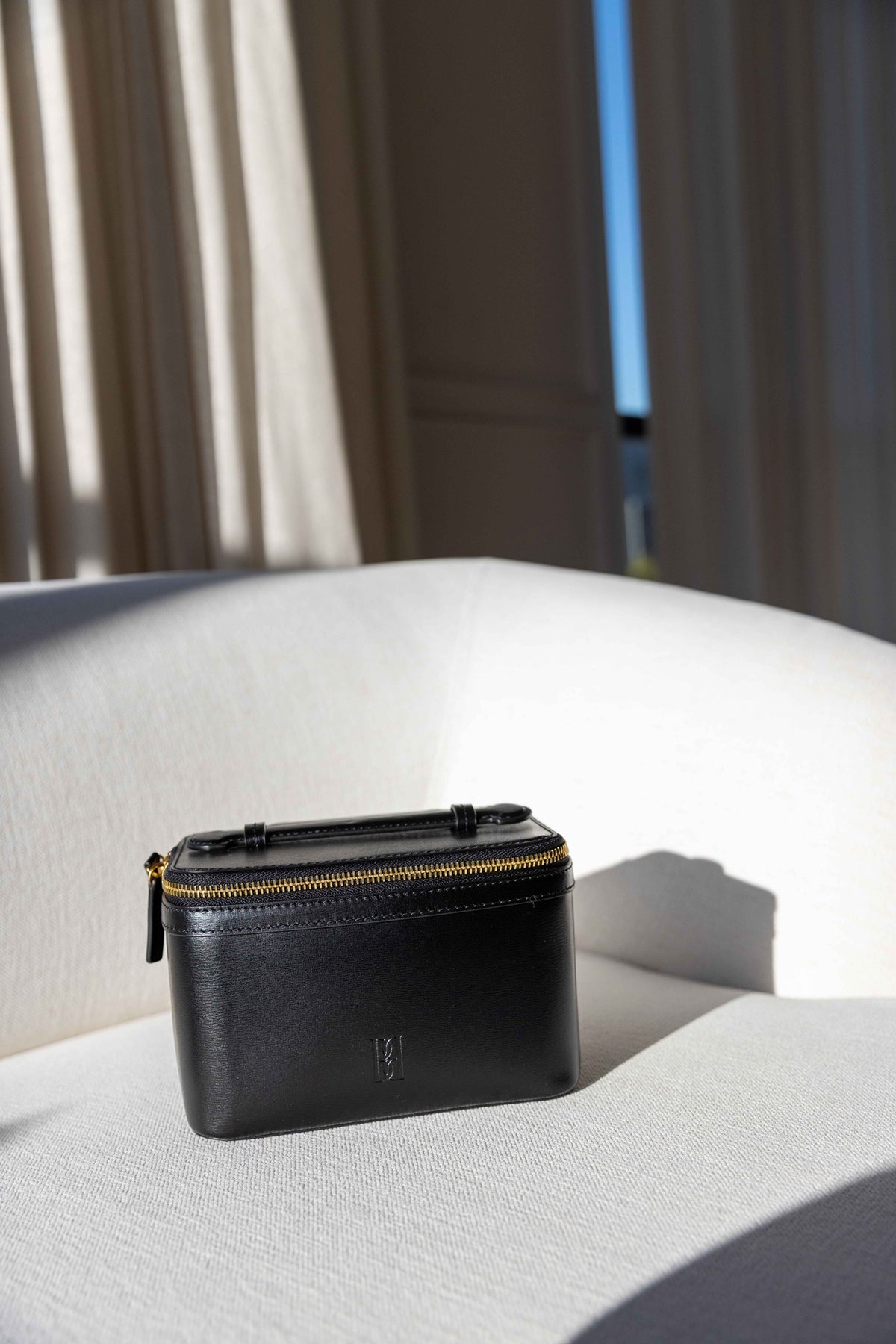 by Malene Birger Aya Beauty Bag Small in black leather, vanity case shape with gold zip, displayed on a white sofa