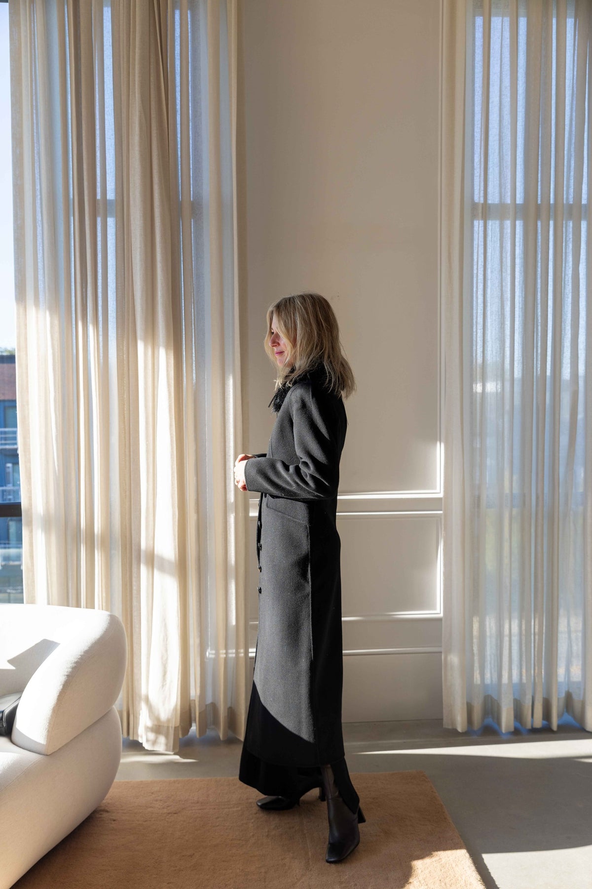 by Malene Birger Magdalena Coat in black, full-length side view of the long tailored wool coat with the Cowie Faux Fur Scarf, in a sunlit room
