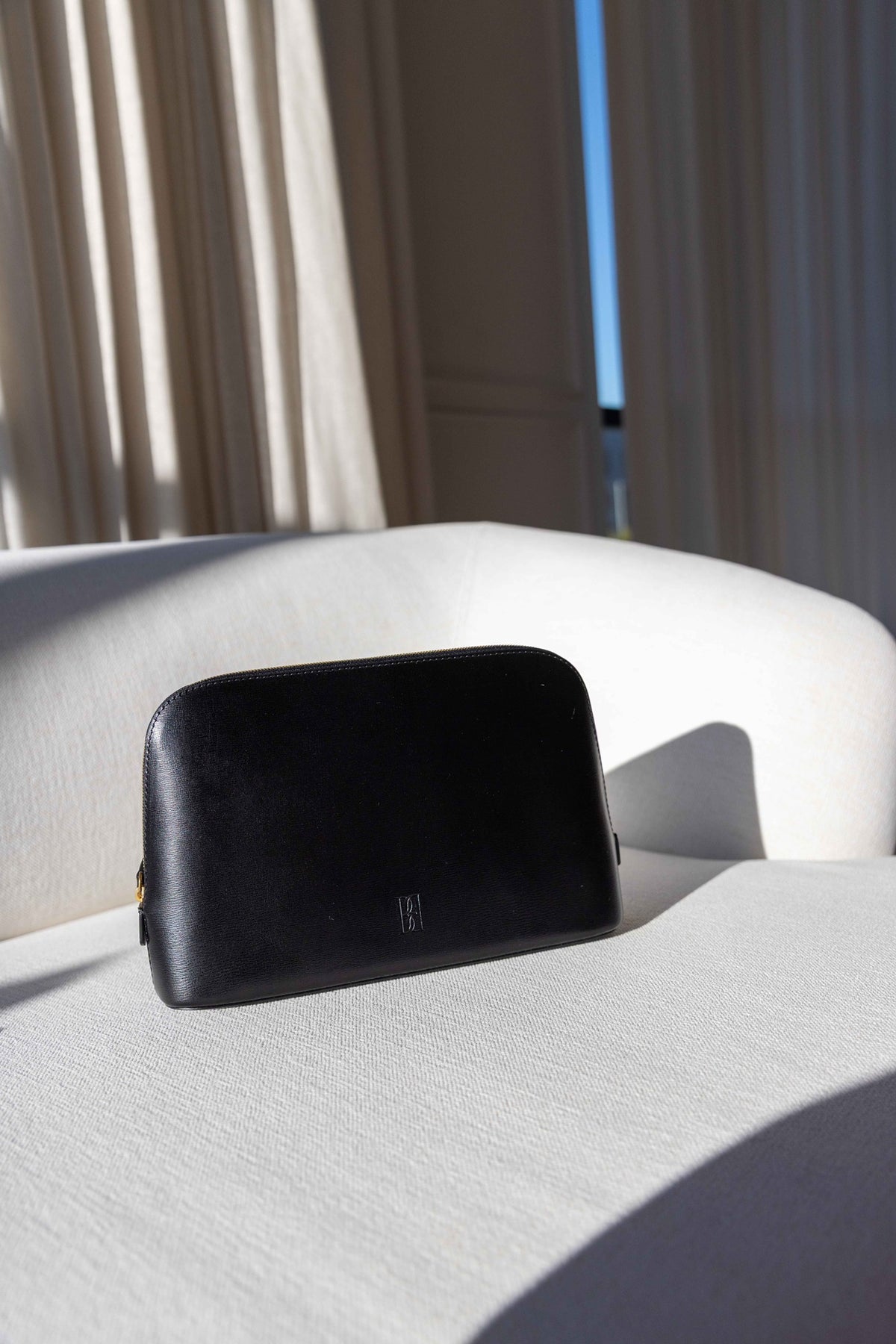 by Malene Birger Aya Medium Cosmetics Case in black leather, domed zip pouch with embossed emblem on a white sofa