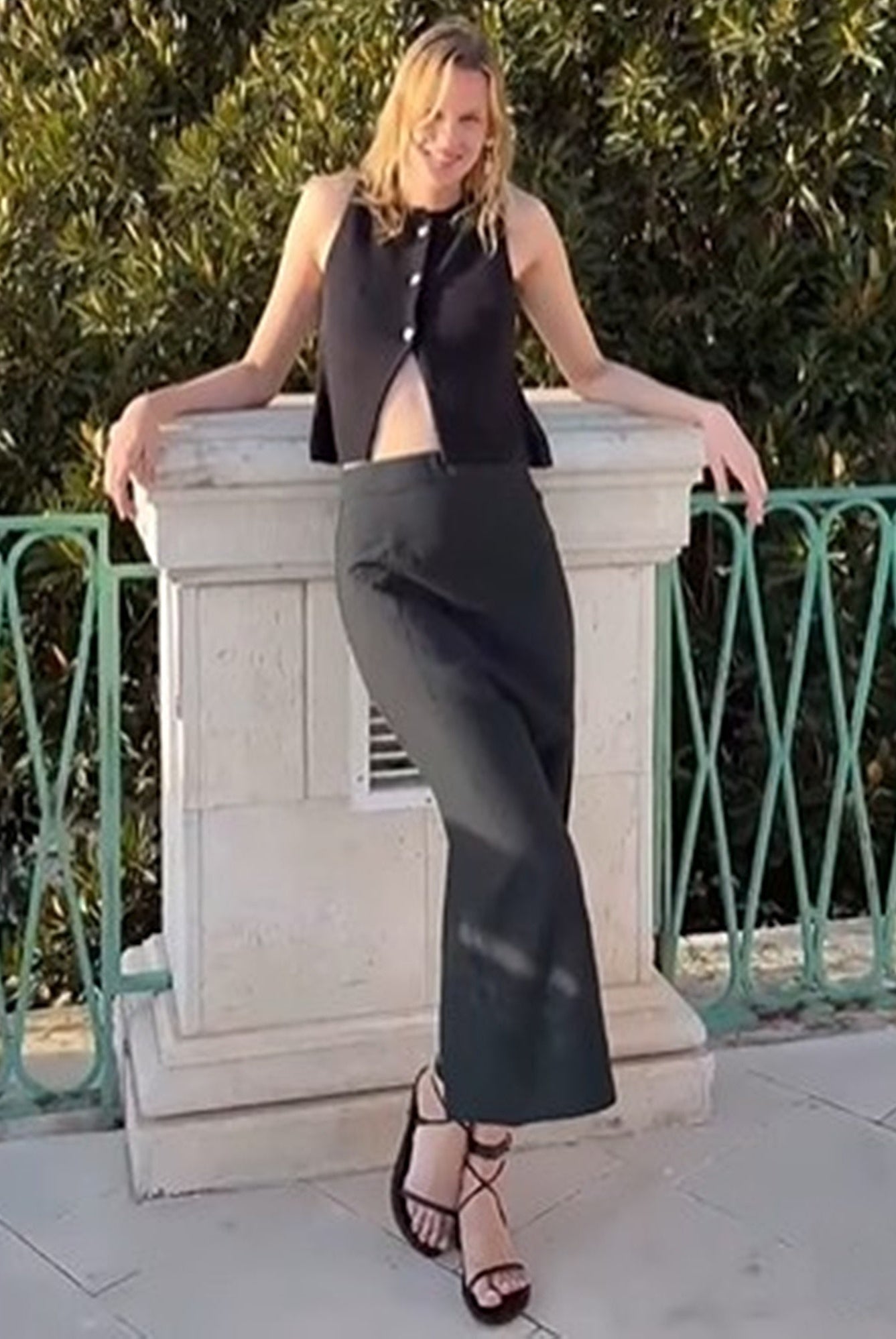 Woman in black sleeveless top and pants leaning against a stone wall with greenery in the background