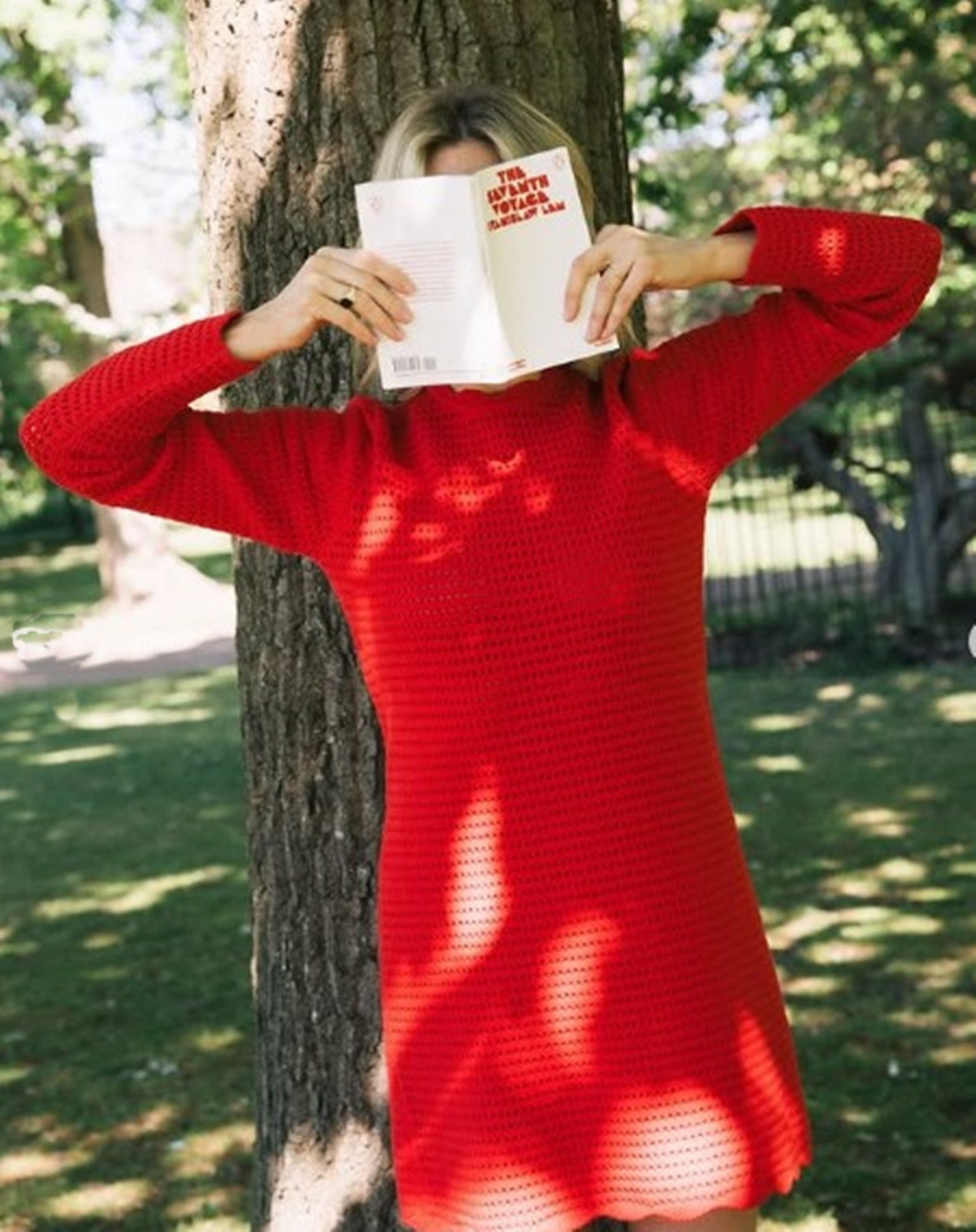 Person in a red dress reading a book outdoors near a tree