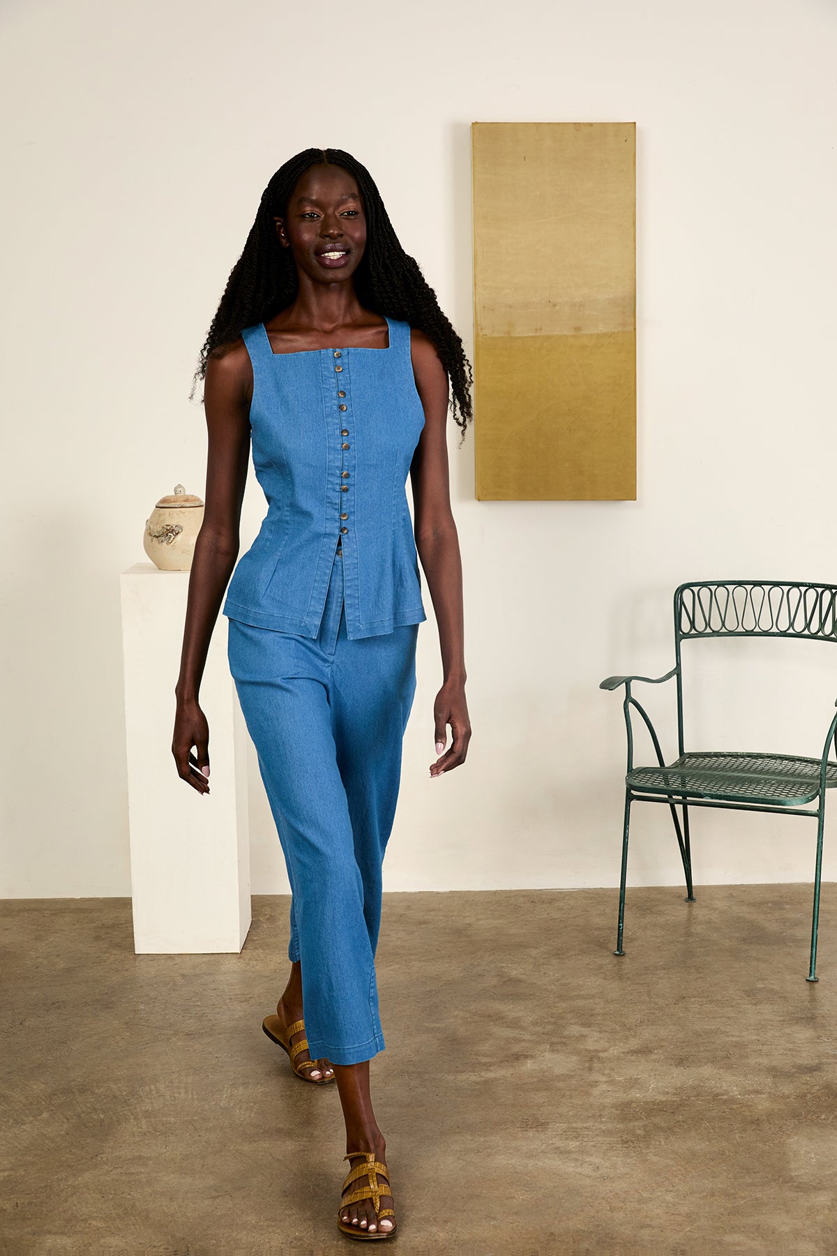 Woman in a blue outfit standing in a room with a chair and wall art.