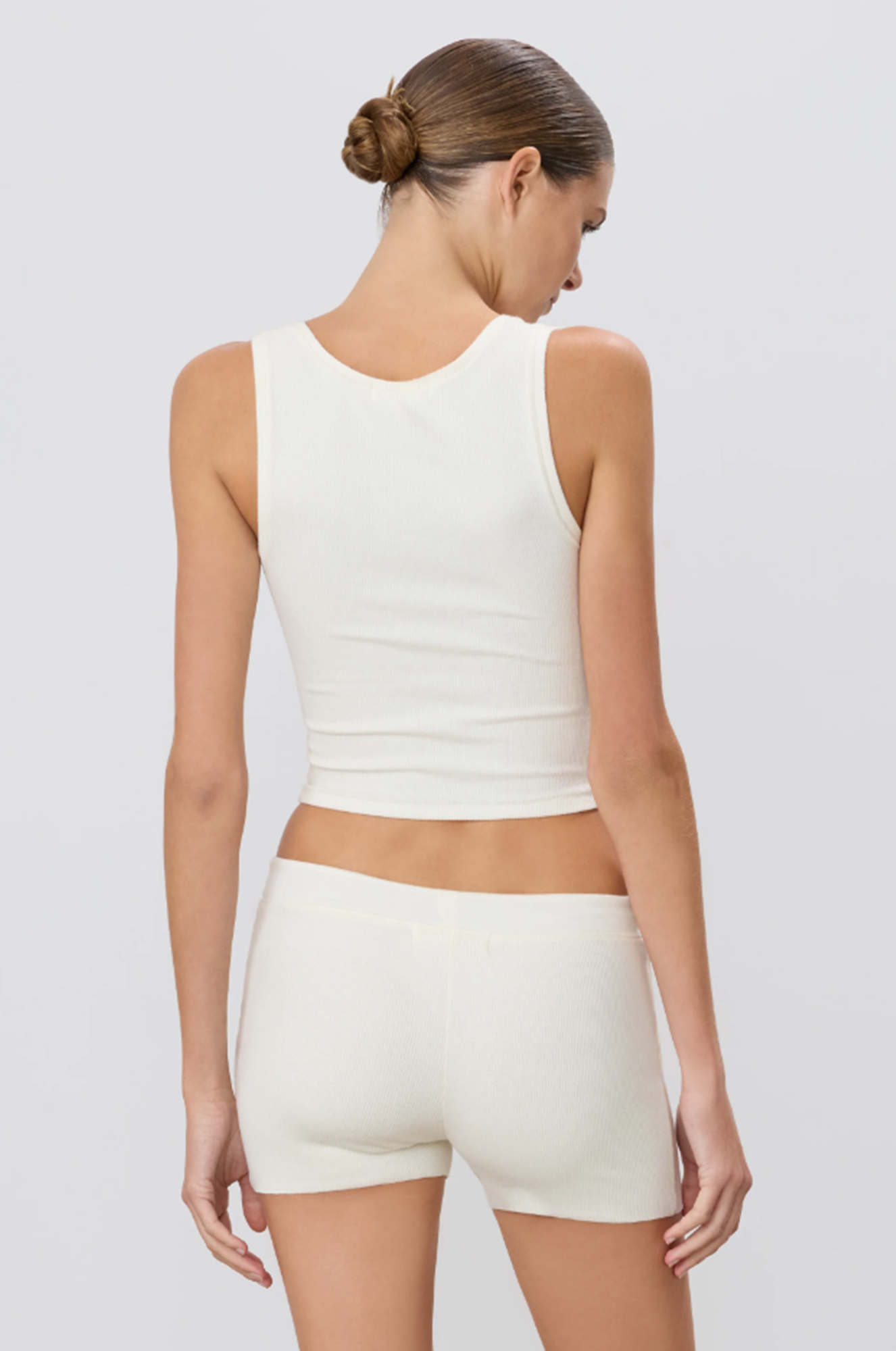 back of a woman wearing a cream sleeveless tank top and shorts on a light gray background