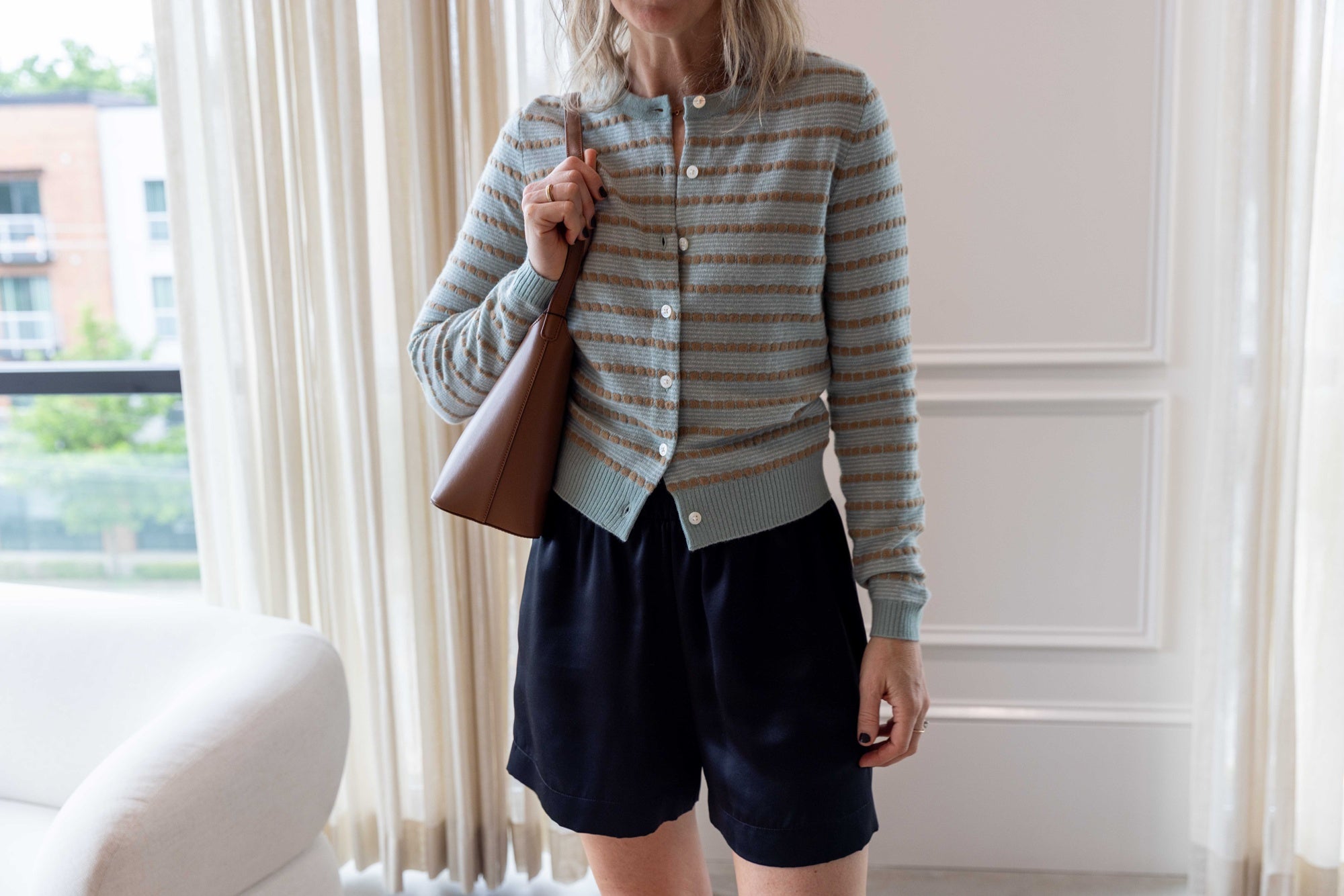 Close up of Woman wearing the Sablyn Brynn Silk Short in black and the guest in residence pebble cardigan carrying an aesther ekme leather handbag standing on a tan rug in front of white curtains.