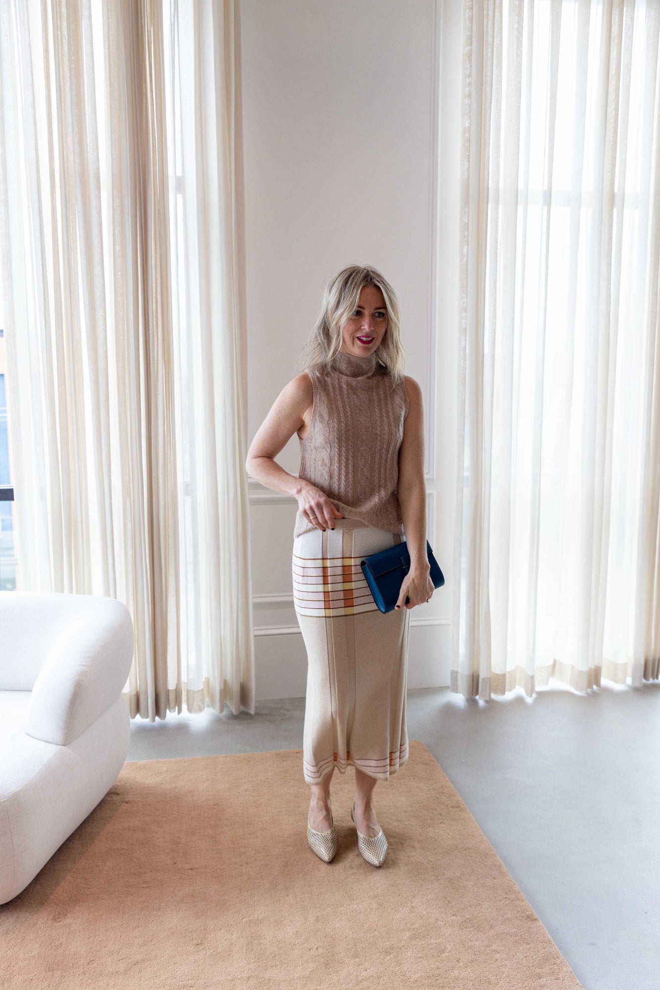 Woman wearing the sablyn salvia sweater and guest in residence plaited rib skirt carrying the hunting season clutch and standing in front of white curtains