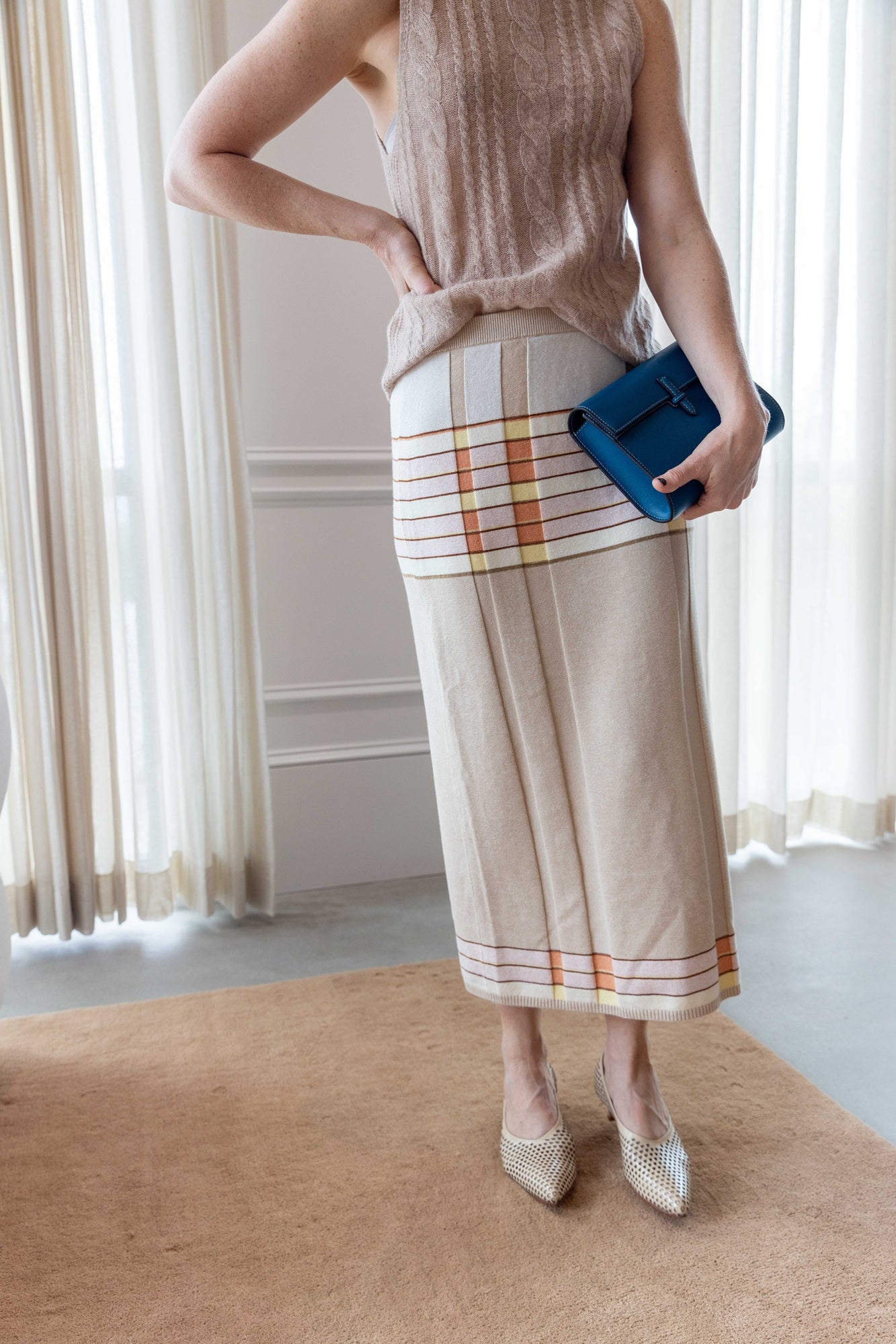 Close up of a Woman wearing the sablyn salvia sweater and guest in residence plaited rib skirt carrying the hunting season clutch and standing in front of white curtains