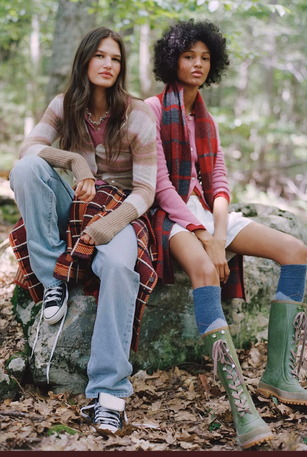 Two women sitting on a rock in a forest, wearing plaid and casual clothing.