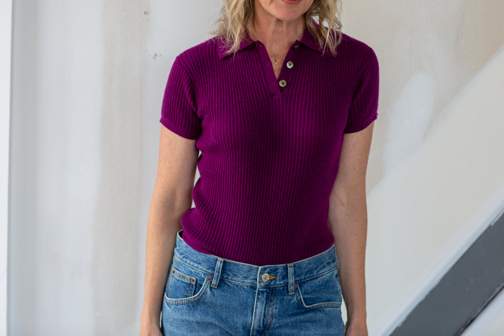 Woman wearing a purple polo shirt and blue jeans against a white wall.