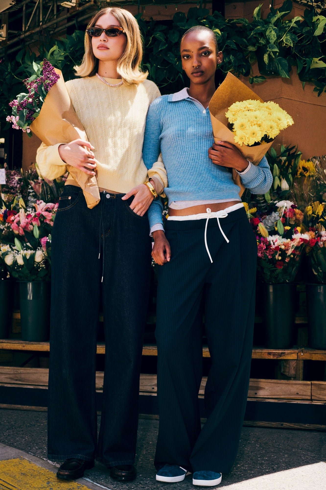 Two women standing together with flowers, wearing stylish outfits in a floral setting.