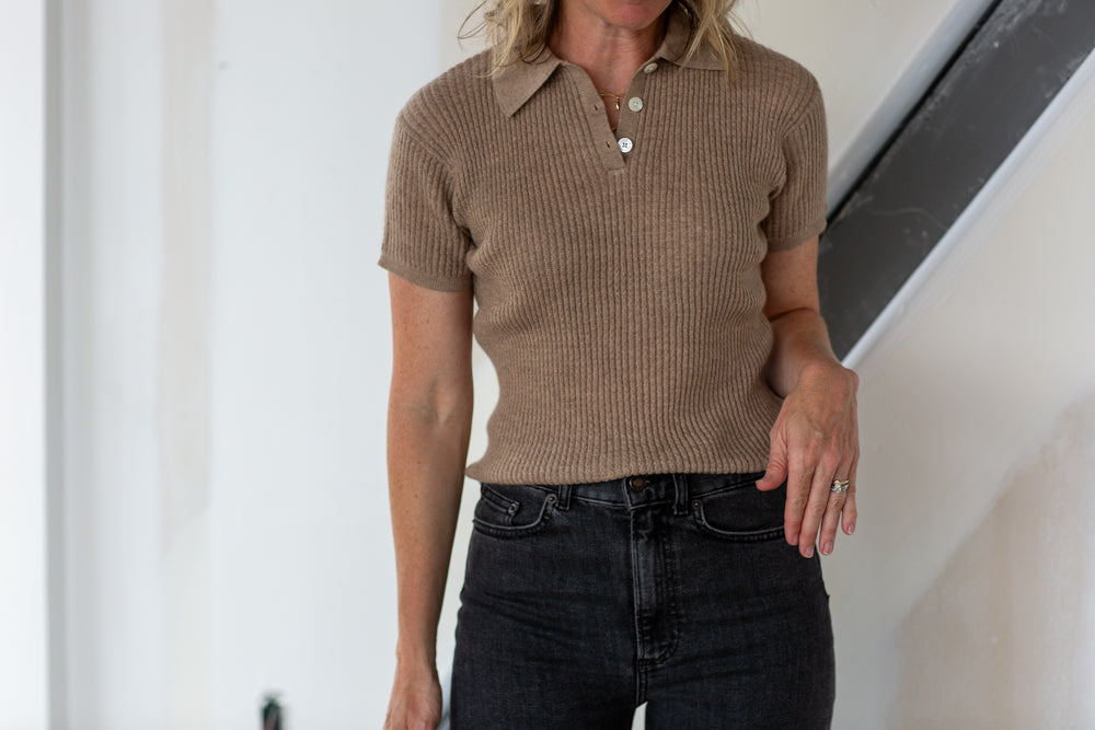 Person wearing a brown ribbed polo shirt and black jeans against a white wall.