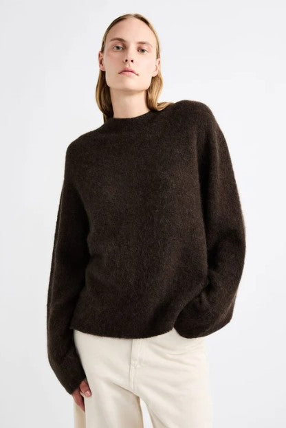 Model wearing the Janessa Leone Laure Sweater in Date, a relaxed dark brown crew-neck knit with cream trousers