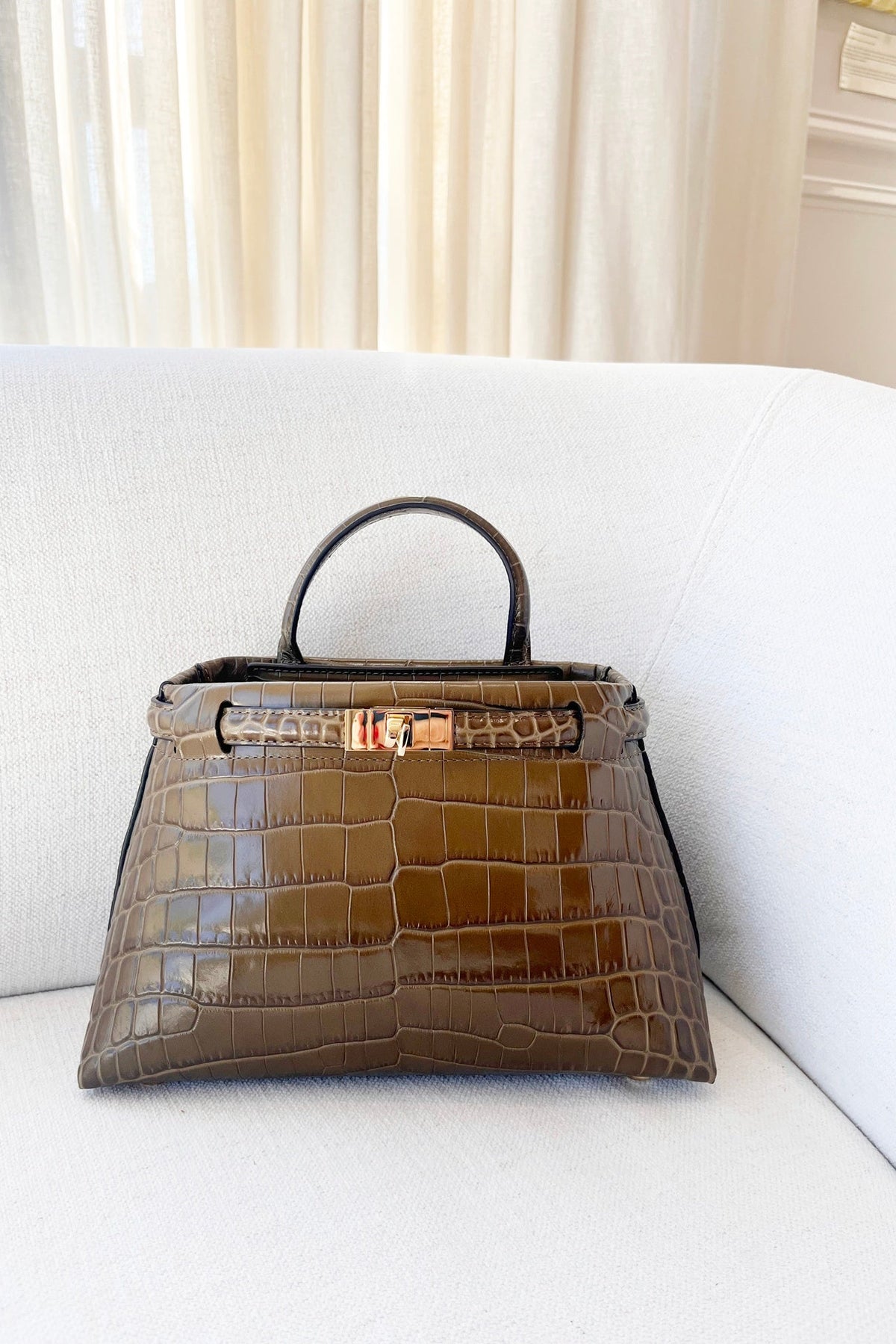 Janessa Leone Mini Leone Bag in Umber Croc, glossy croc-embossed leather top-handle bag with gold-tone clasp, front view on a cream sofa