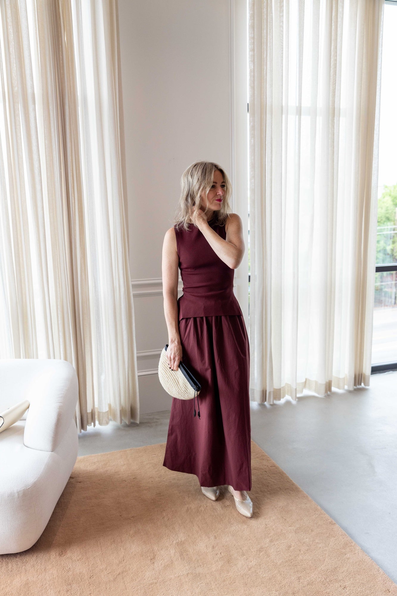 Woman wearing the Onia boatneck dress in dark cherry holding the hunting season clutch and standing on a tan rug in front of white curtains.