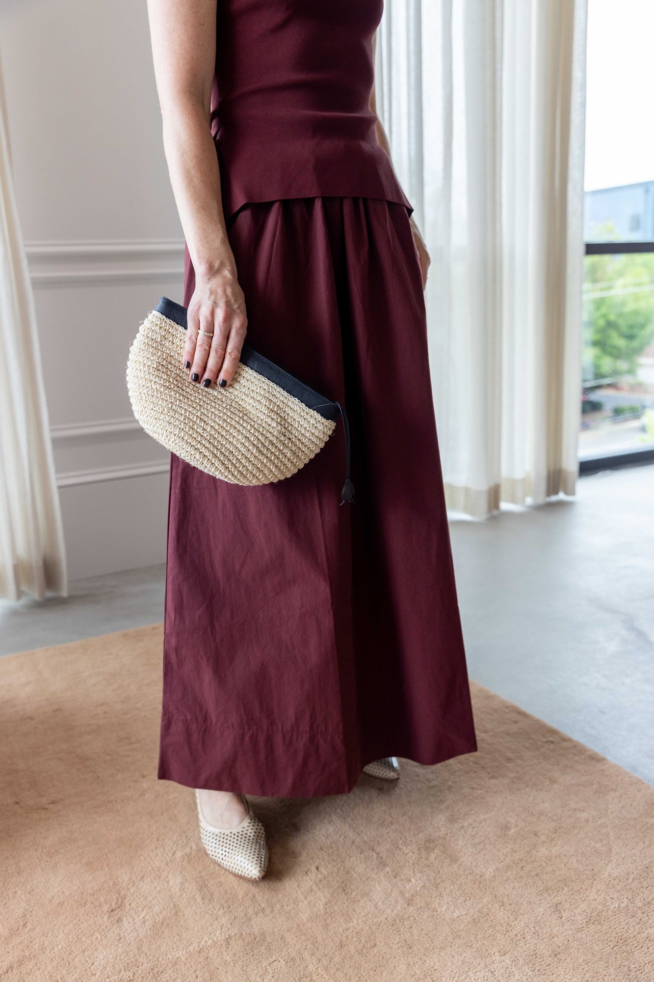 Close up of a Woman wearing the Onia boatneck dress in dark cherry holding the hunting season clutch and standing on a tan rug in front of white curtains.