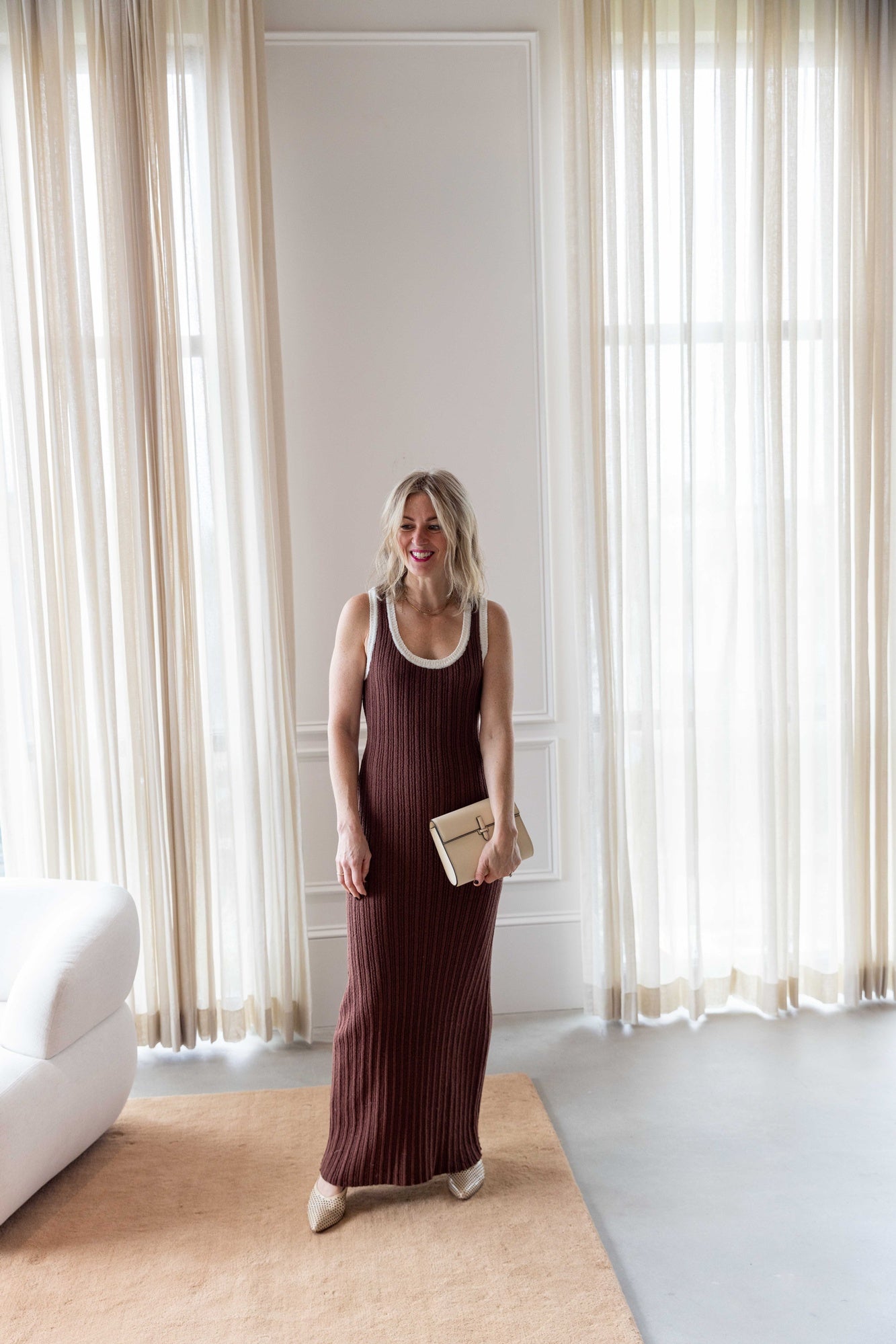 Woman wearing the Onia Cotton Boucle Scoop Tank Dress and sablyn lance sweater carrying a hunting season clutch standing on a tan carpet in front of white curtains.