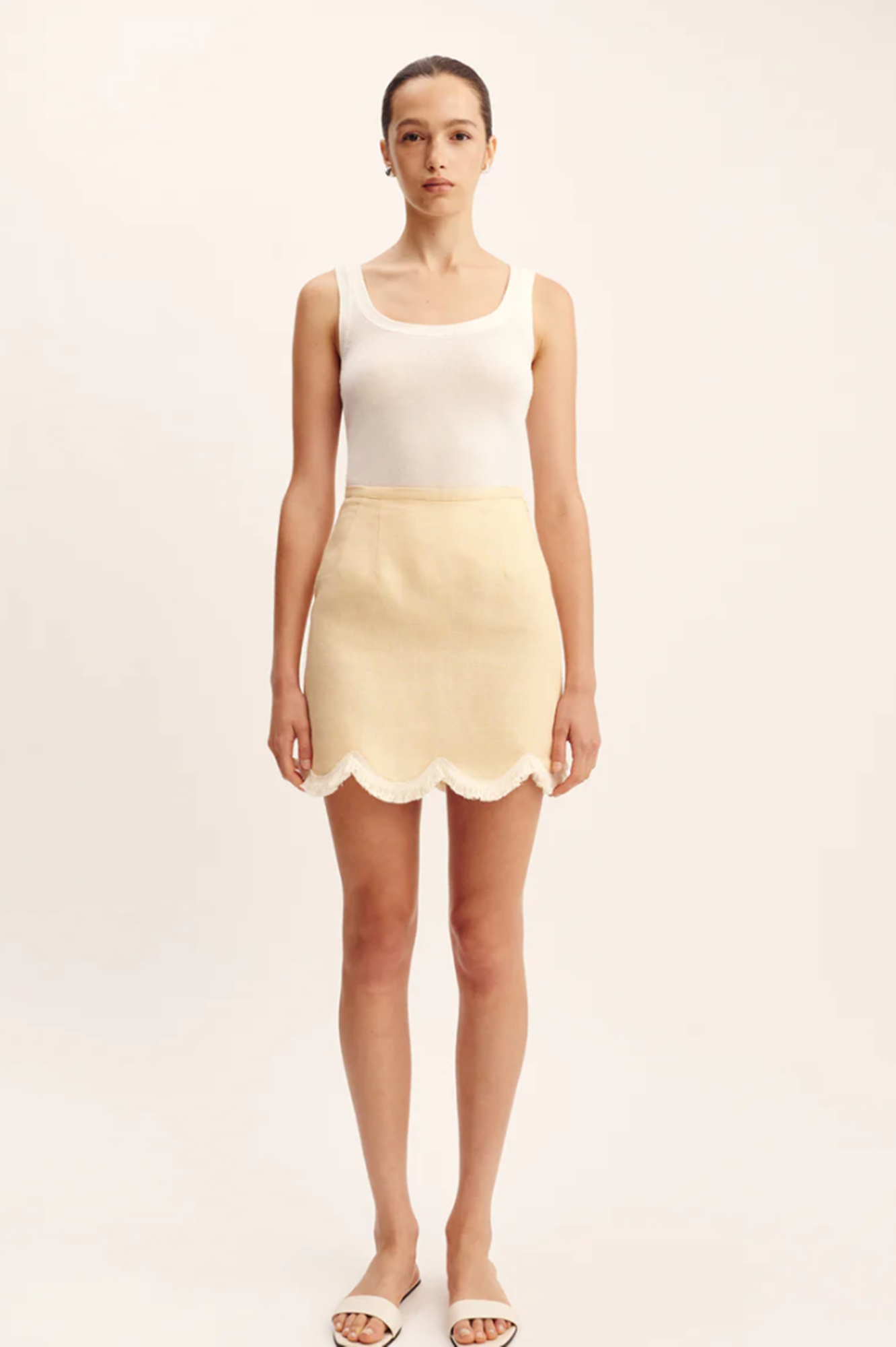 Woman wearing a white tank top and beige skirt with scalloped edges on a plain background