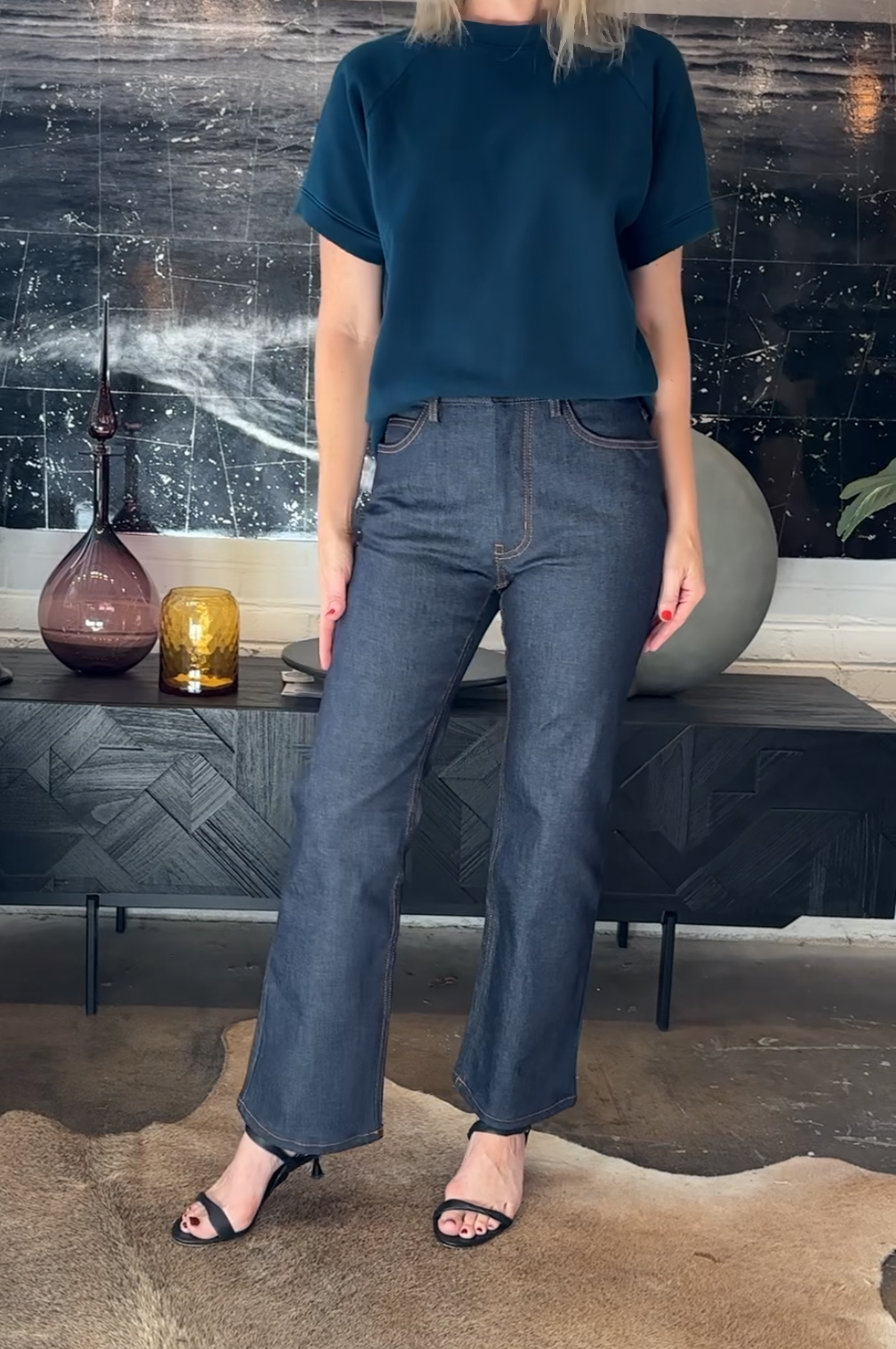 Person wearing a blue top and jeans standing in a room with decorative items on a shelf.