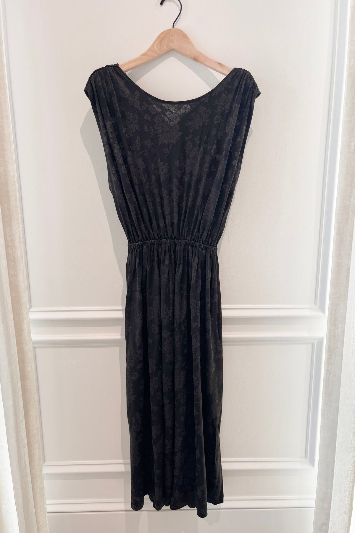 A sleeveless black midi dress with a textured, slightly sheer lace-like bodice and an elasticized waist, hanging on a wooden hanger against a white paneled wall.