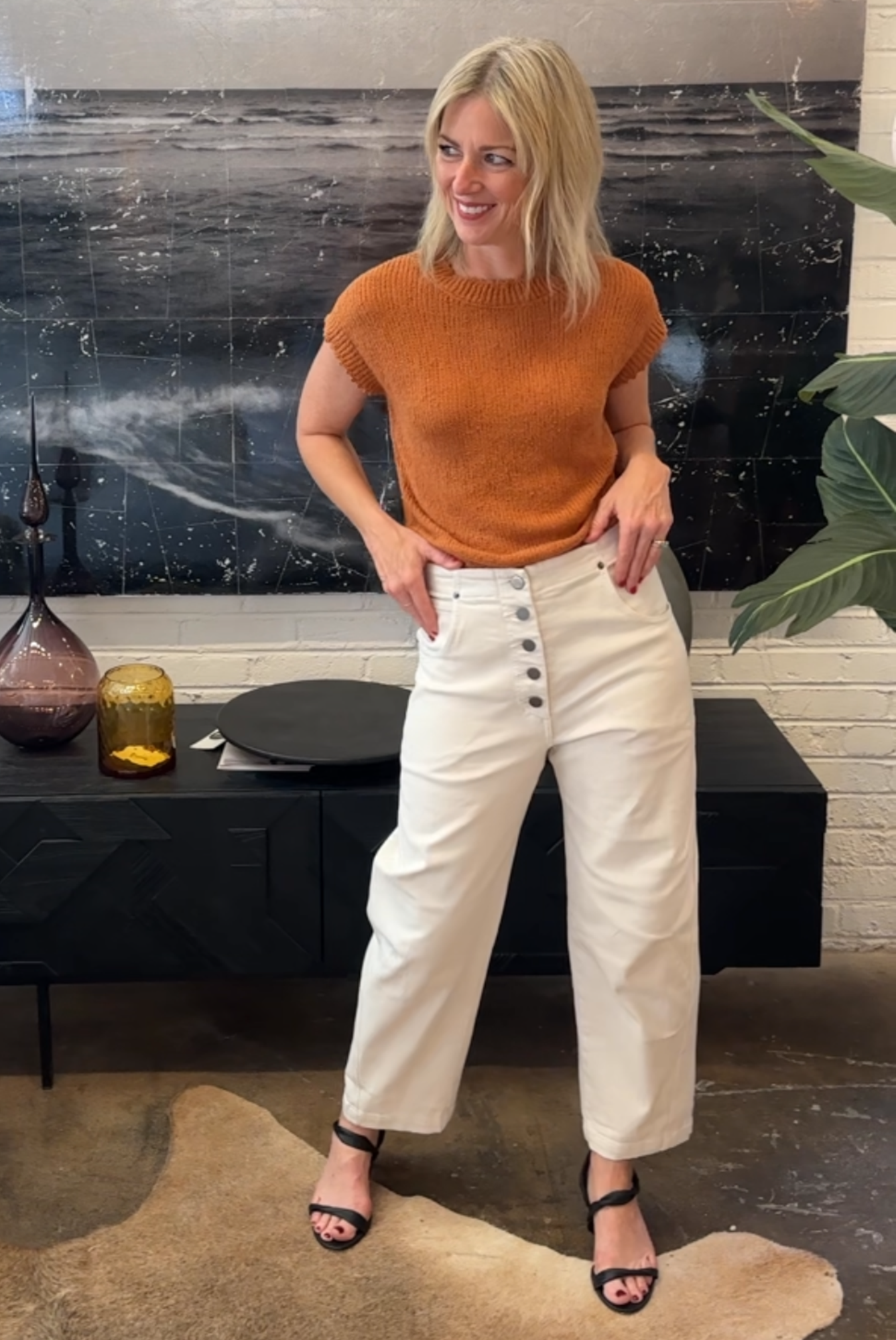 Woman in an orange top and white pants standing in a modern living room.