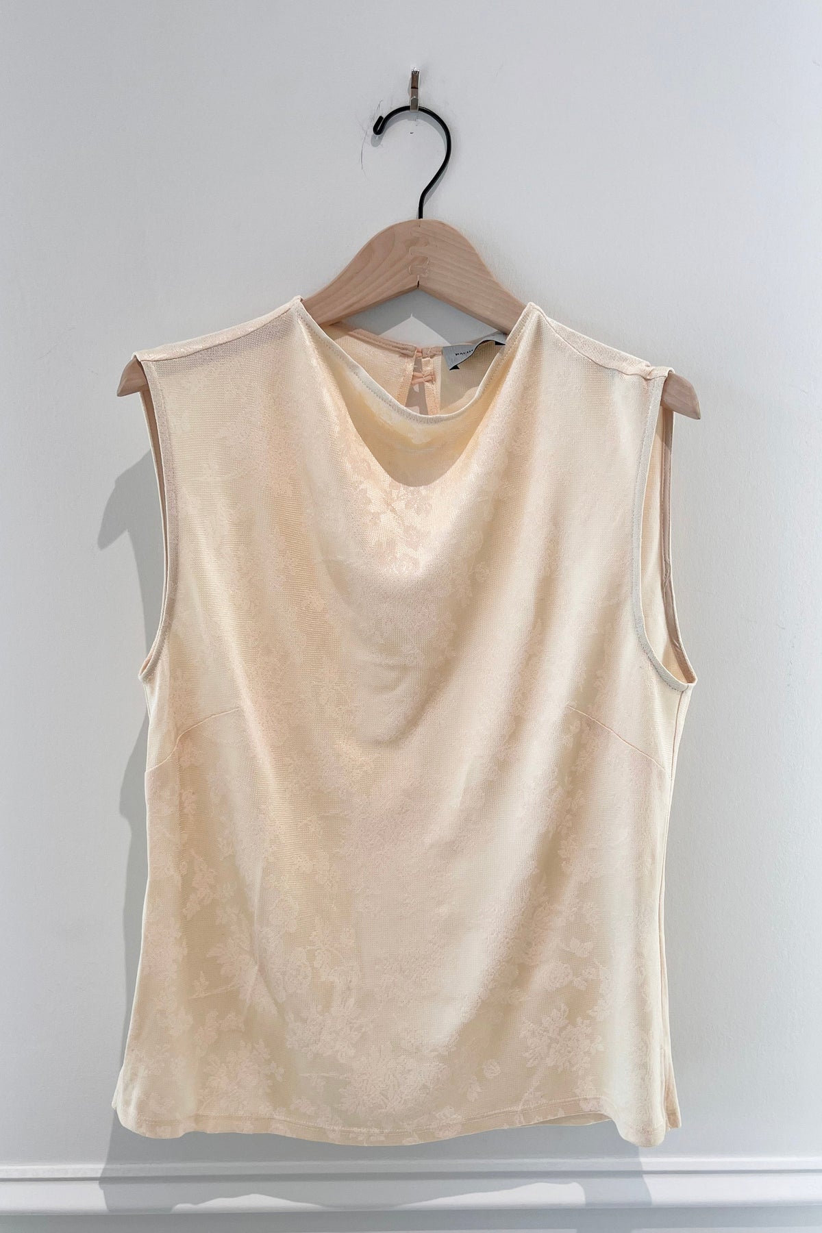 A sleeveless, scoop-neck top in a light cream or beige color, hanging on a wooden hanger against a plain light gray wall.