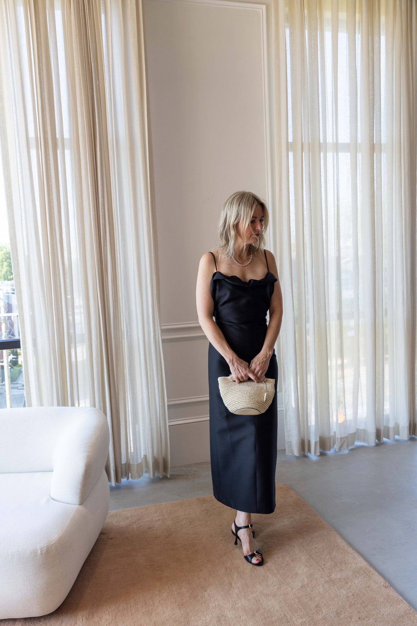 A woman wearing the Rachel Comey Priss Dress and holding a hunting season handbag standing on a tan rug in front of white curtains