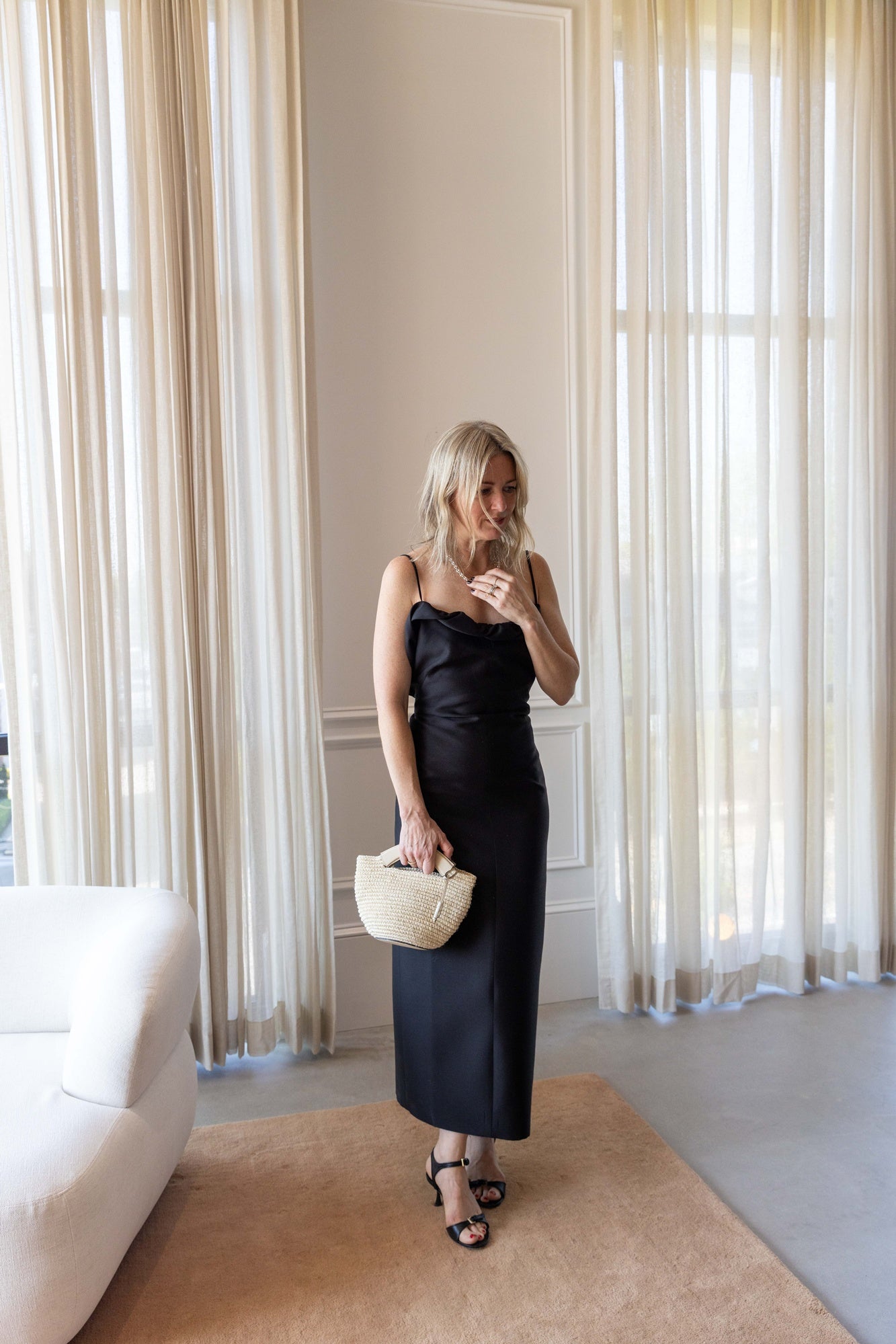 A woman wearing the Rachel Comey Priss Dress and holding a hunting season handbag standing on a tan rug in front of white curtains