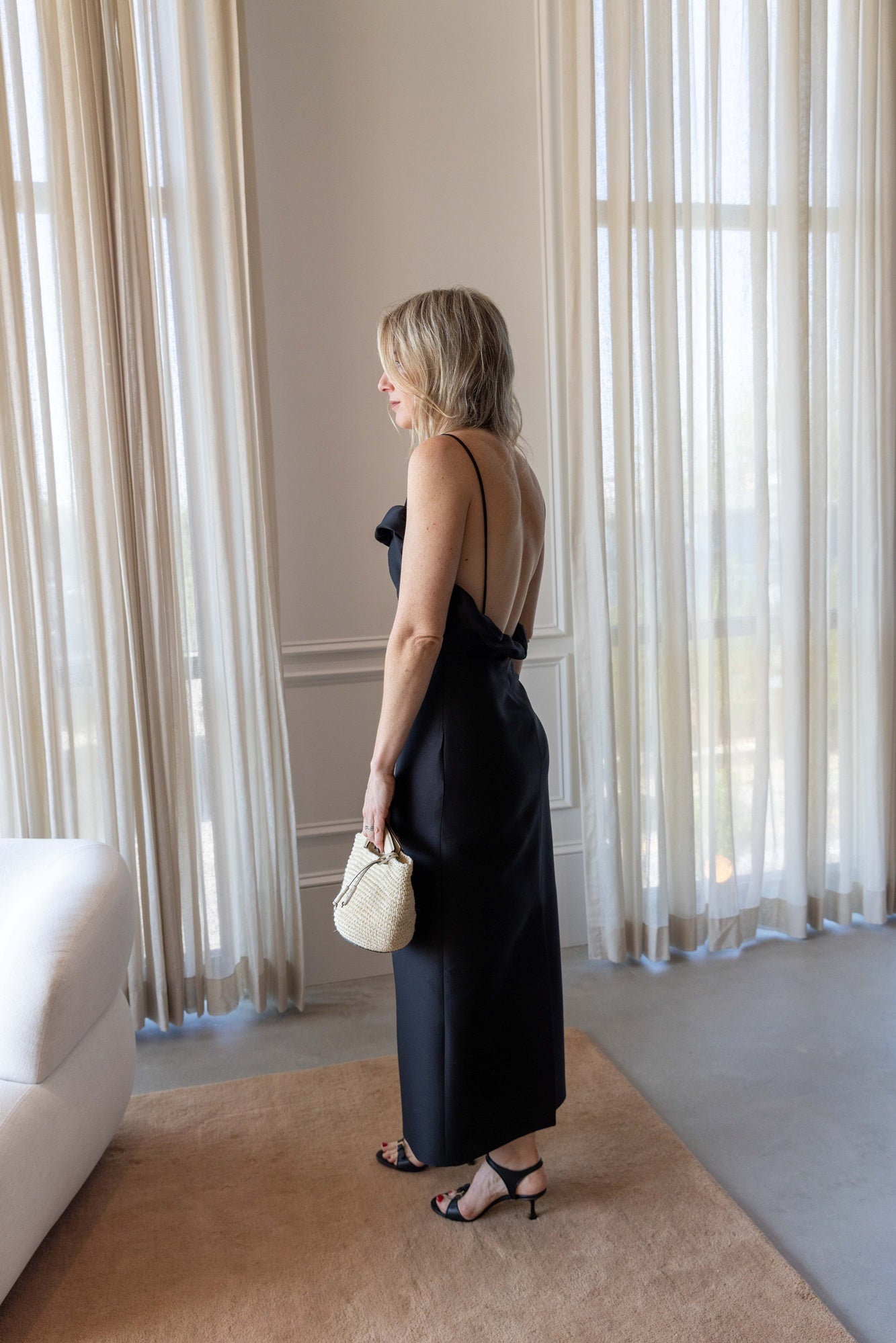 Backside of a woman wearing the Rachel Comey Priss Dress and holding a hunting season handbag standing on a tan rug in front of white curtains