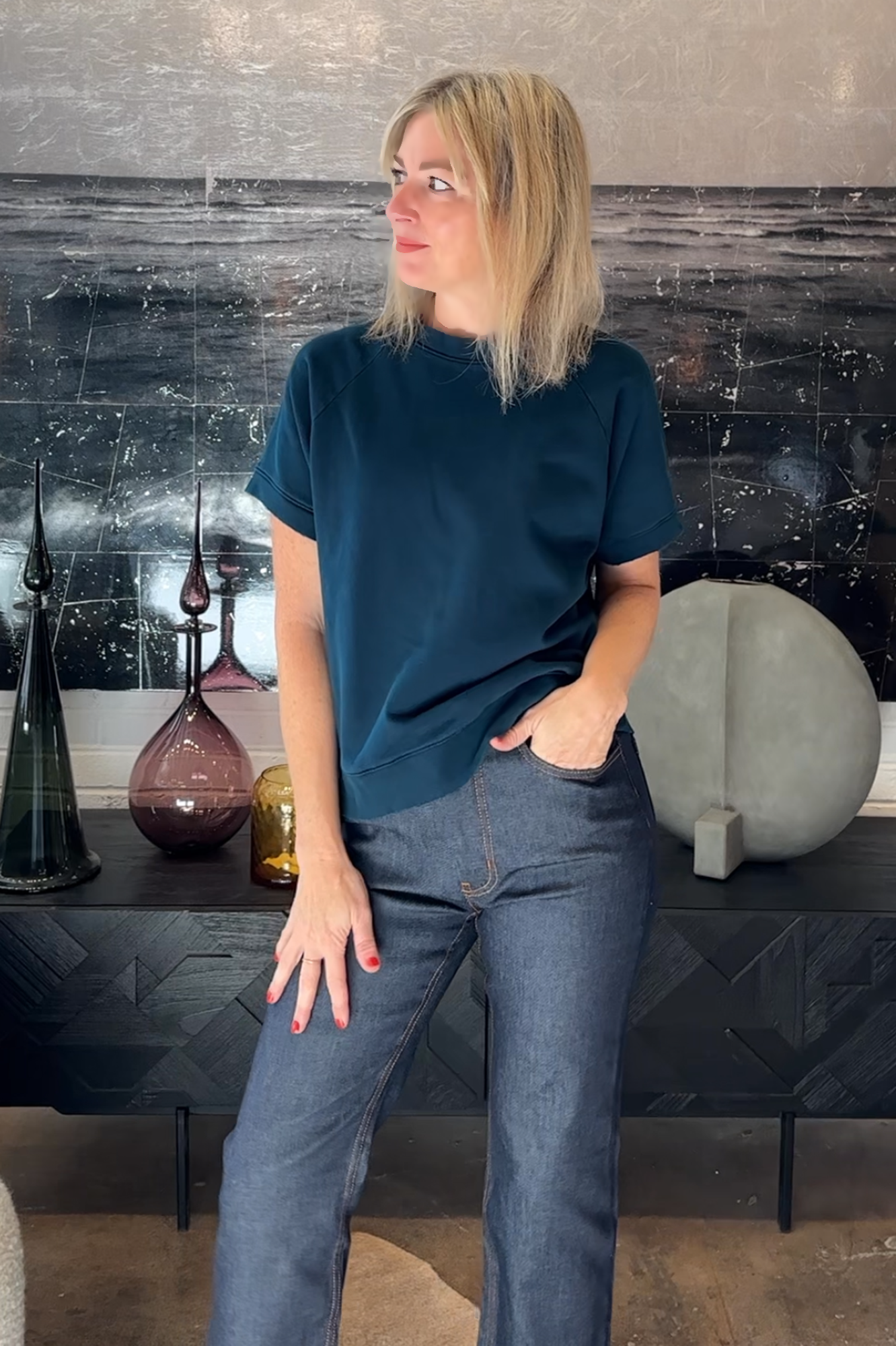 Woman in a teal shirt and jeans standing in a room with decorative vases and a textured wall.