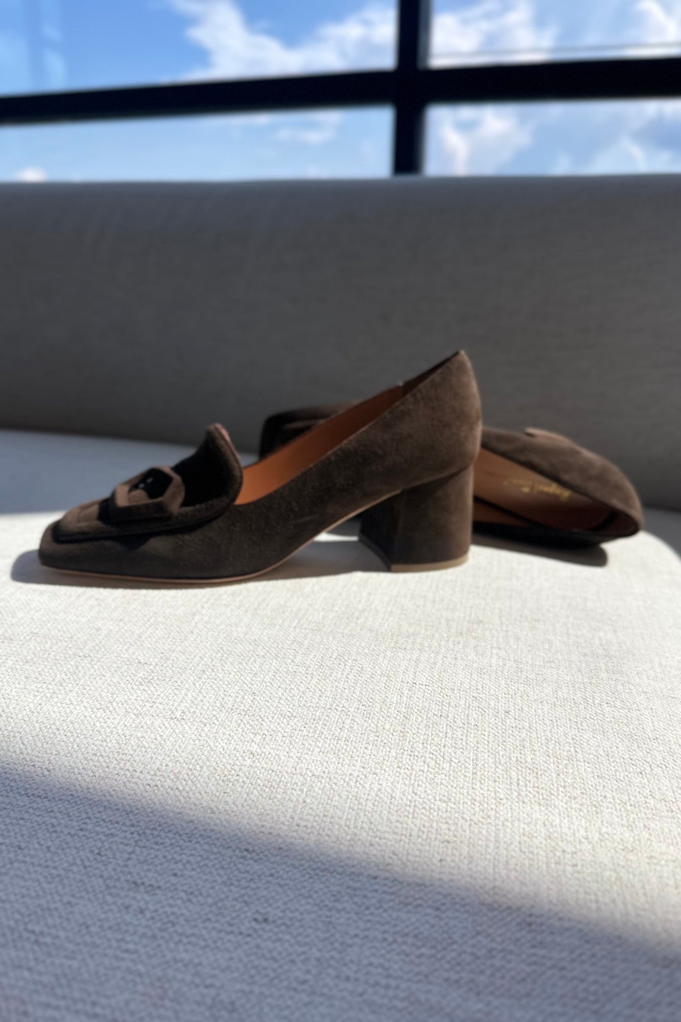 Brown suede shoes with a block heel on a textured surface with a window in the background