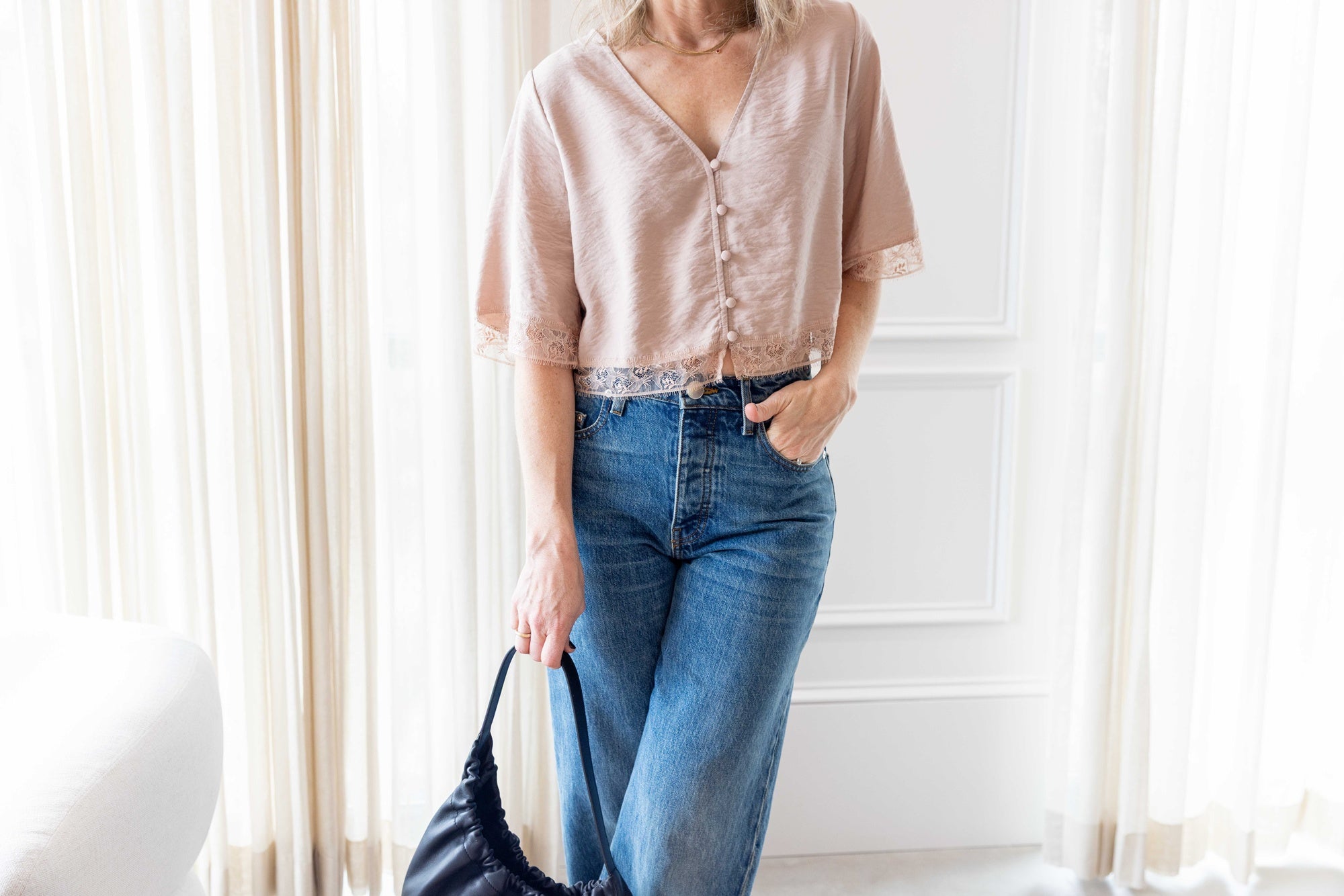Close up of a woman wearing the Sablyn Simone SS Button Down Top with Lace in dune and the sammy jeans carrying the aesther ekme handbag standing in front of white curtains