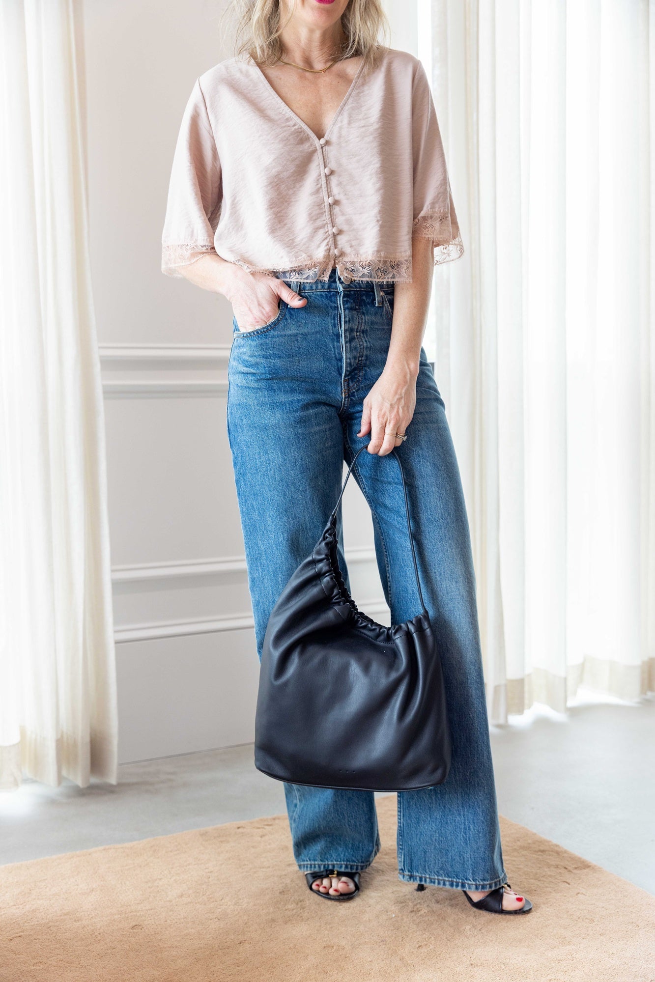 Woman wearing the Sablyn Simone SS Button Down Top with Lace in dune and the sammy jeans carrying the aesther ekme handbag standing in front of white curtains