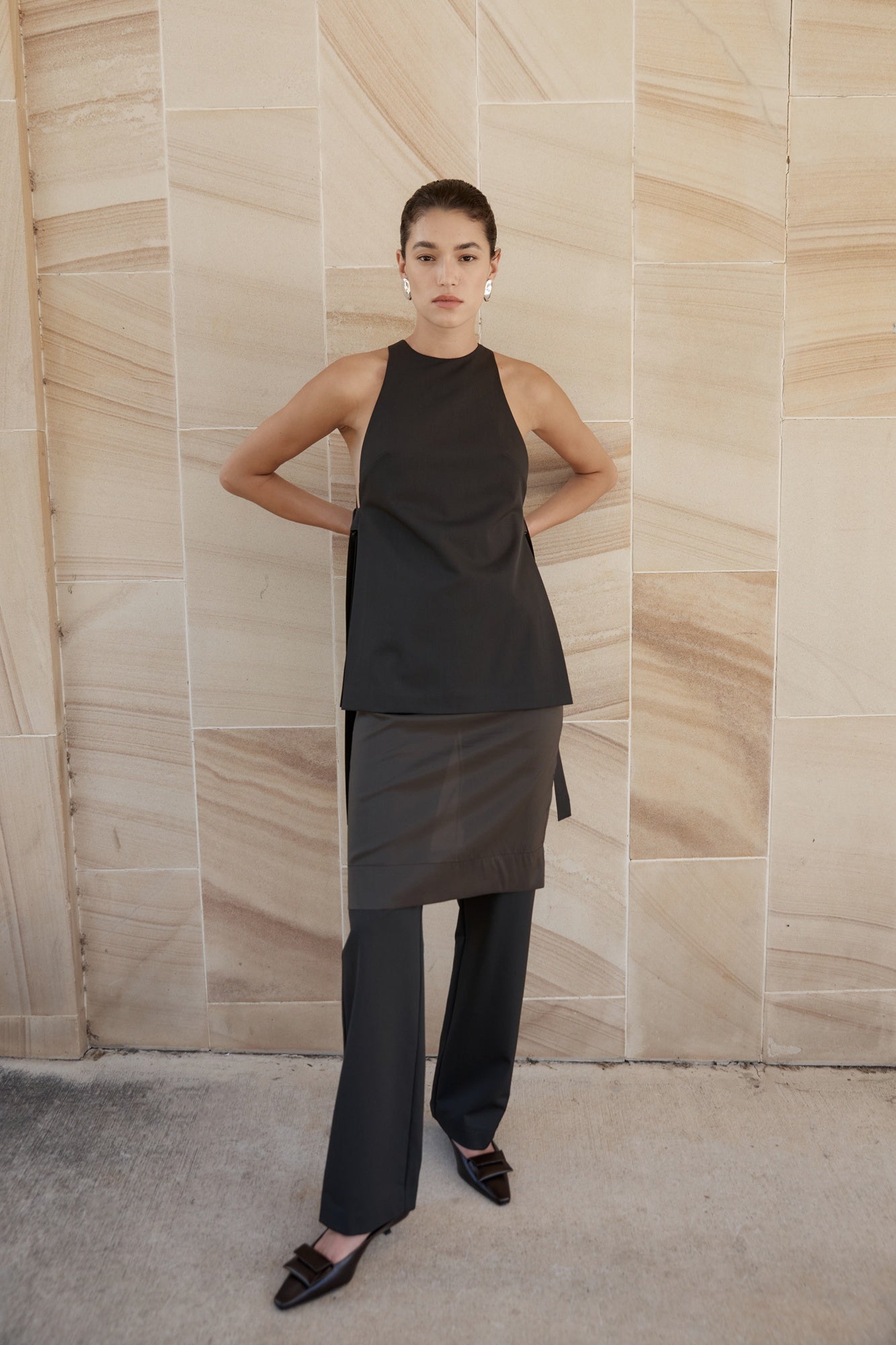 Woman wearing a black sleeveless top and dark pants against a beige stone wall.