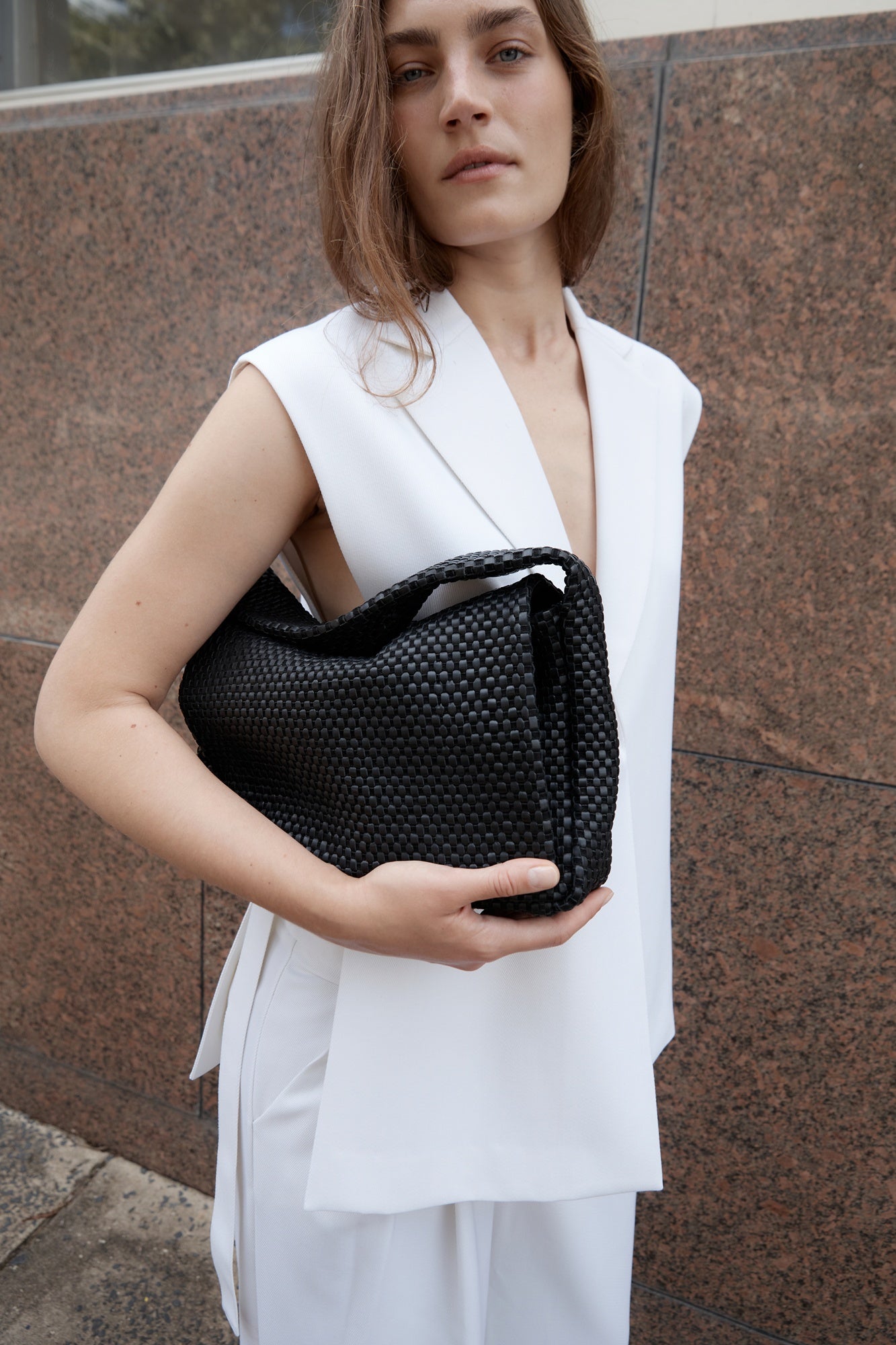 Woman holding a black woven handbag against a brown wall.