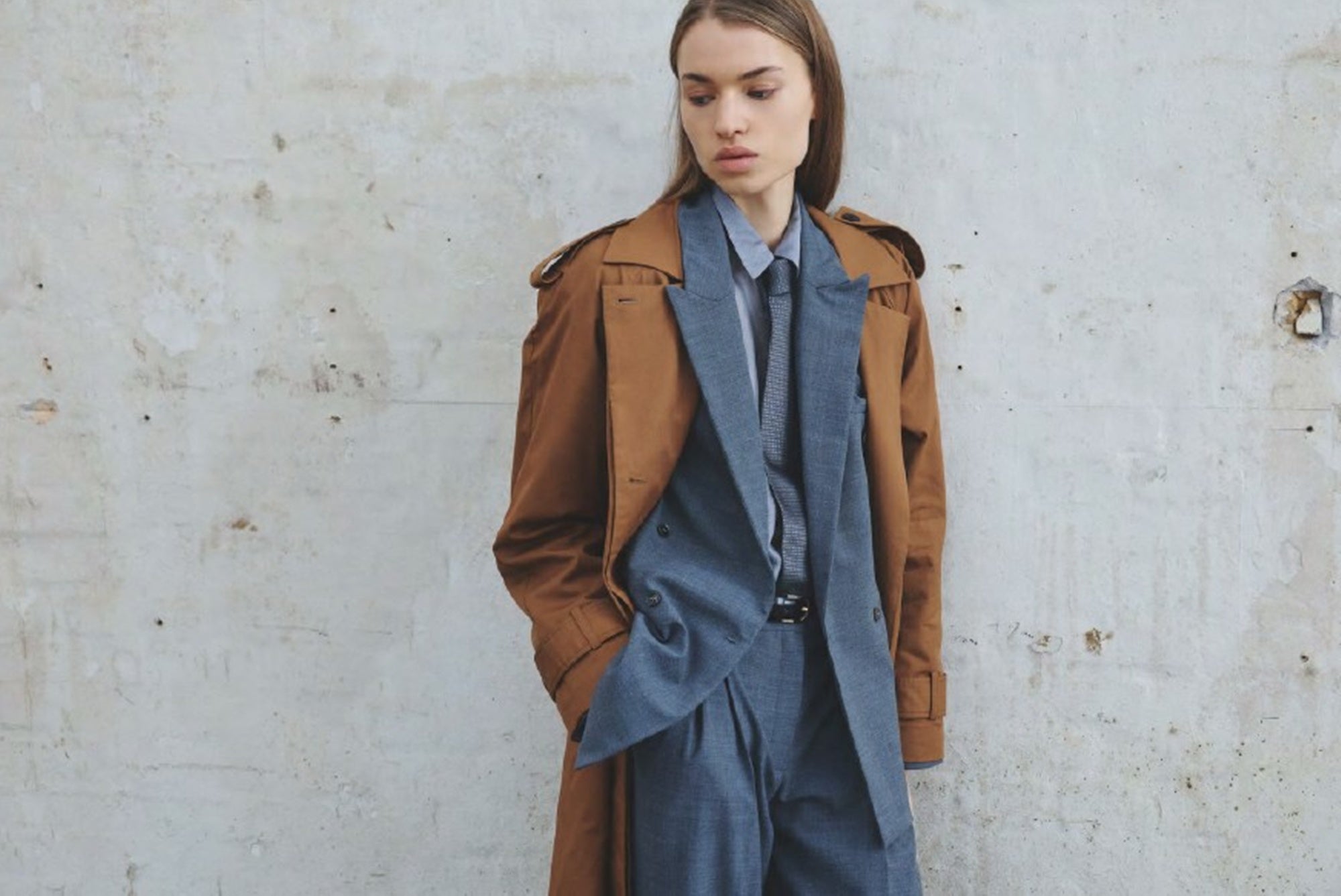 Woman wearing a brown coat over a blue outfit against a light gray wall.