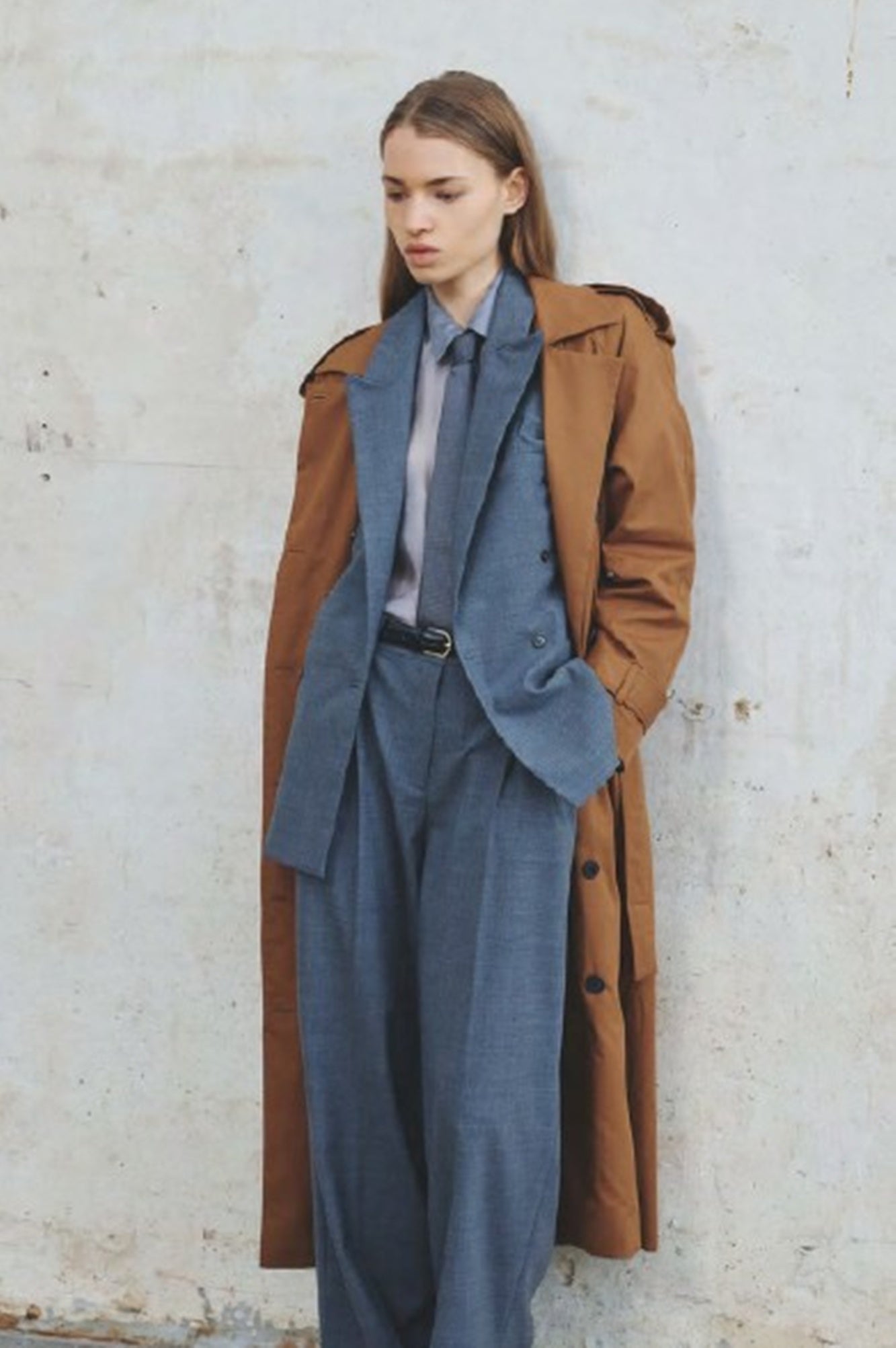 Person wearing a long brown coat over a blue suit against a light-colored wall.